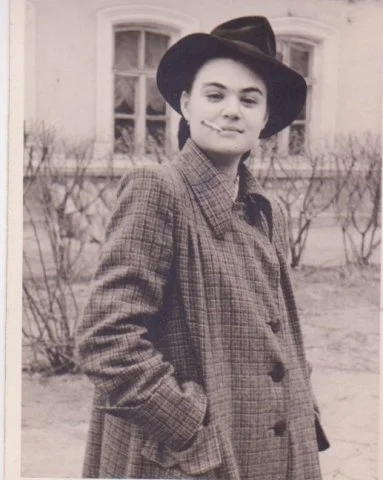 An archival photograph of a woman in a brimmed hat and checked coat standing outdoors.