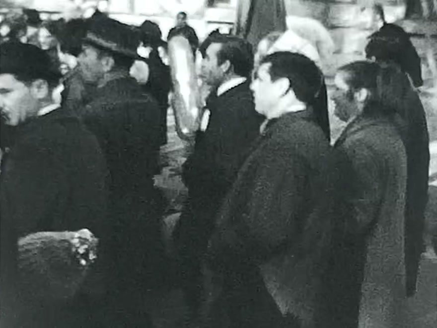 Black-and-white home-movie footage of adults and children in coats moving through an outdoor crowd with balloons.