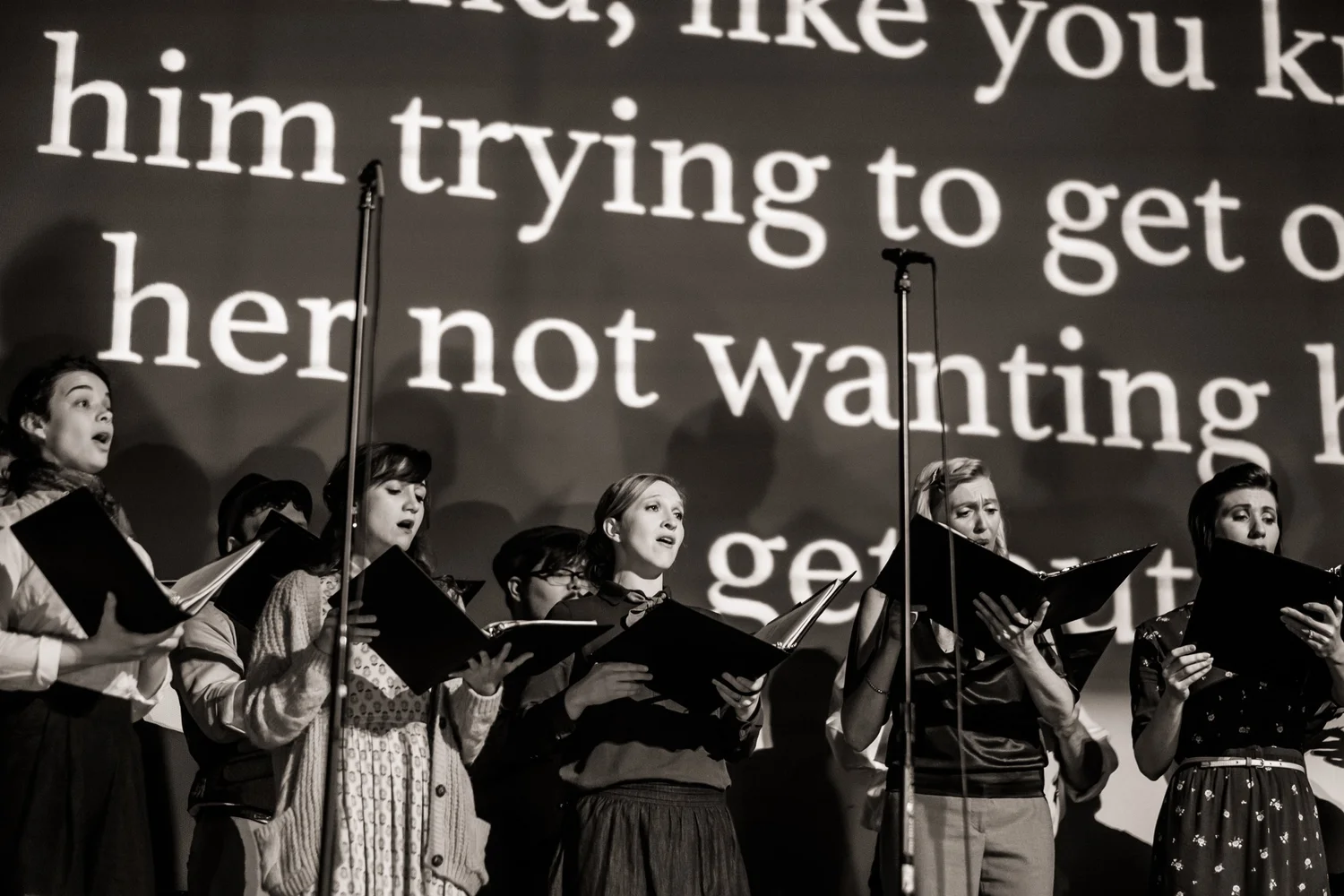 Verdigris Ensemble singers before enlarged fragments of the radio transcript in an archival 2019 performance of Betty’s Notebook.
