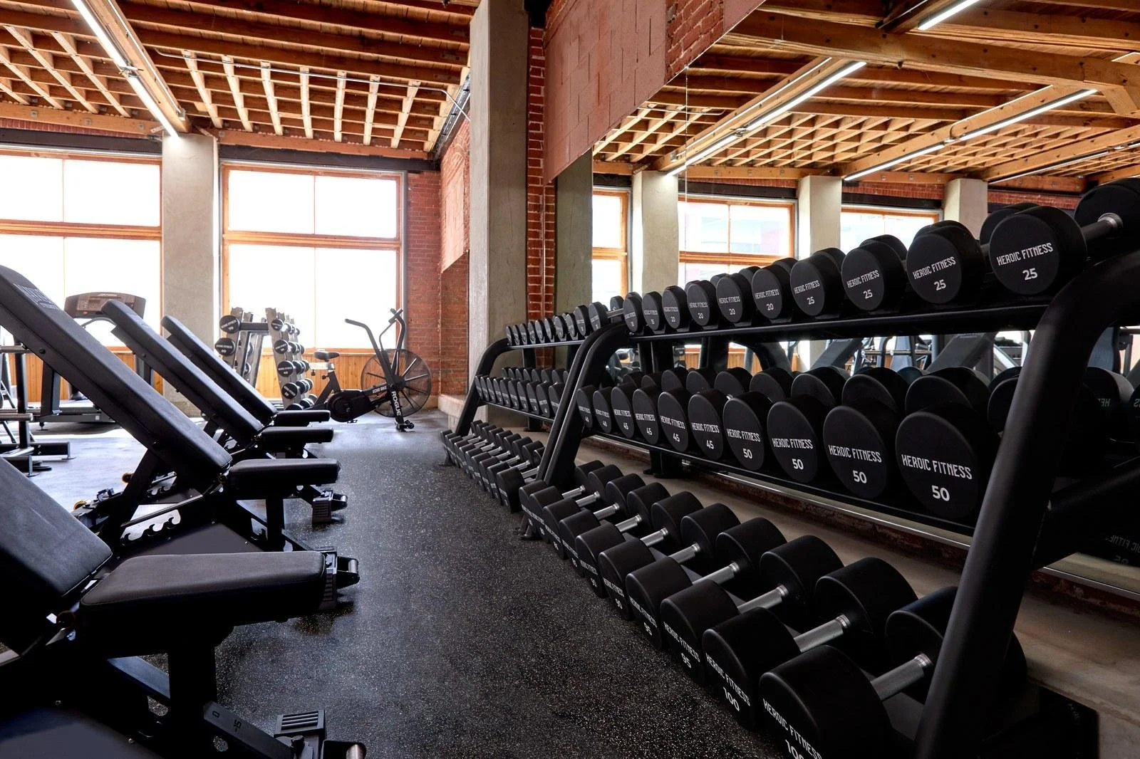 Strength floor with dumbbell racks and benches under timber beams and tall windows