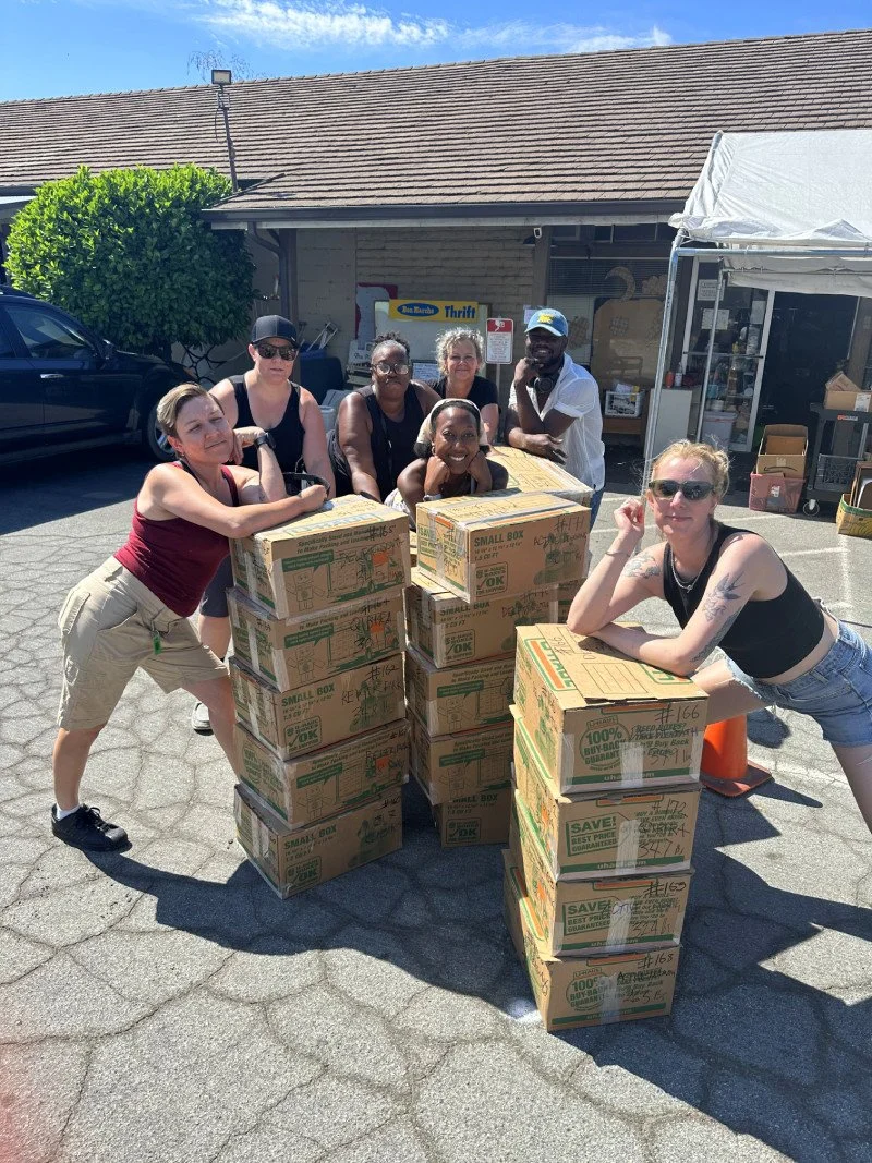 A diverse group of seven COSAS volunteers stand around a stack of boxes packed and ready for shipping