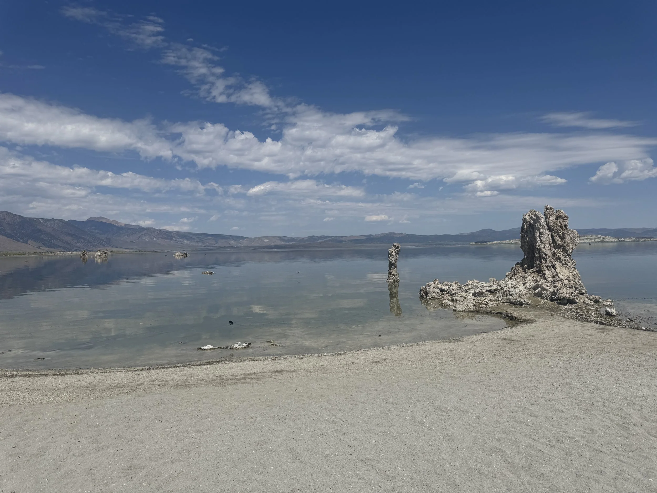 Tufa towers rising from the calm water of Mono Lake beneath dramatic white clouds and a deep blue sky.