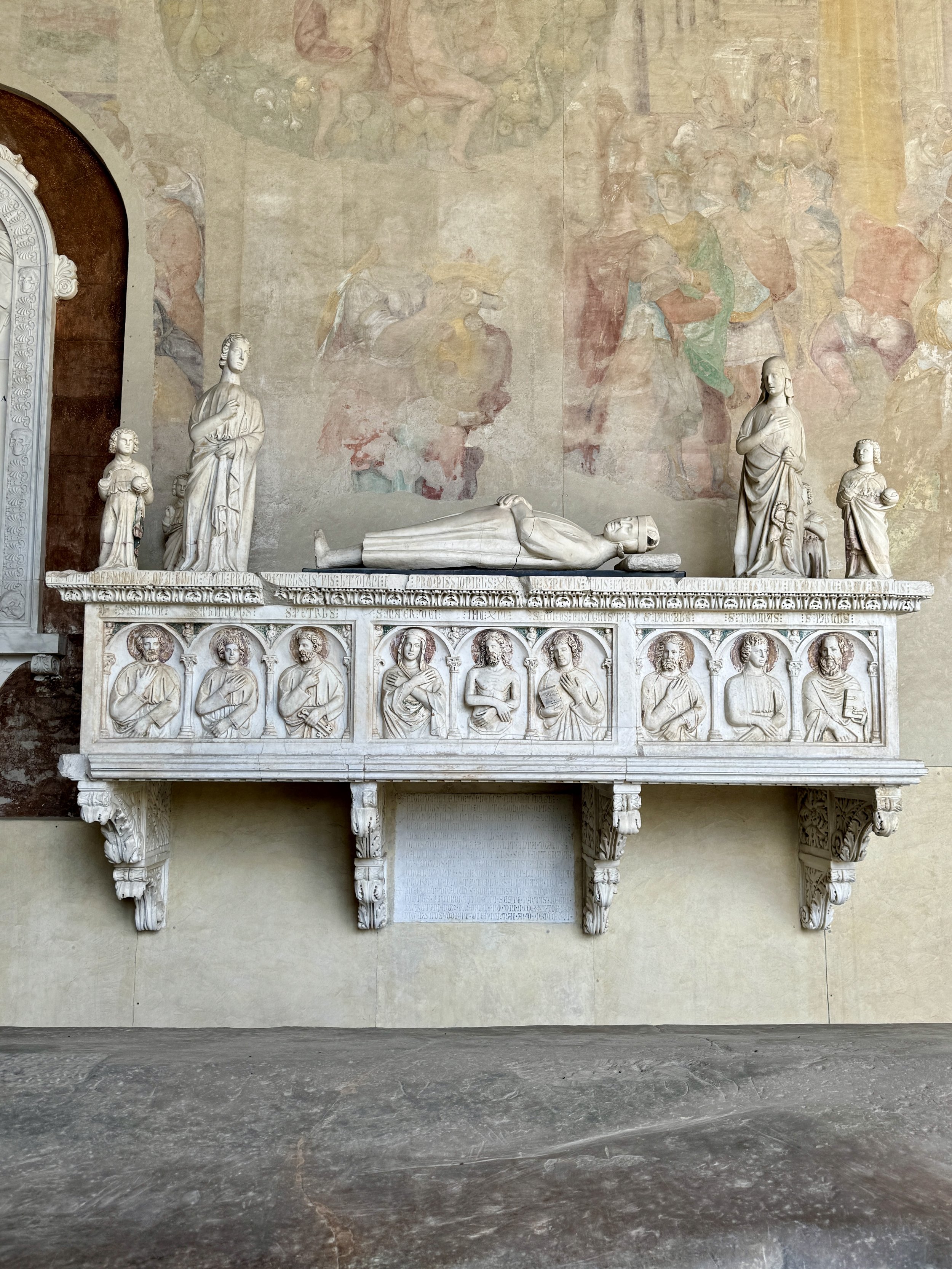 Ornate wall tomb with reclining effigy and saint reliefs inside Camposanto Monumentale.