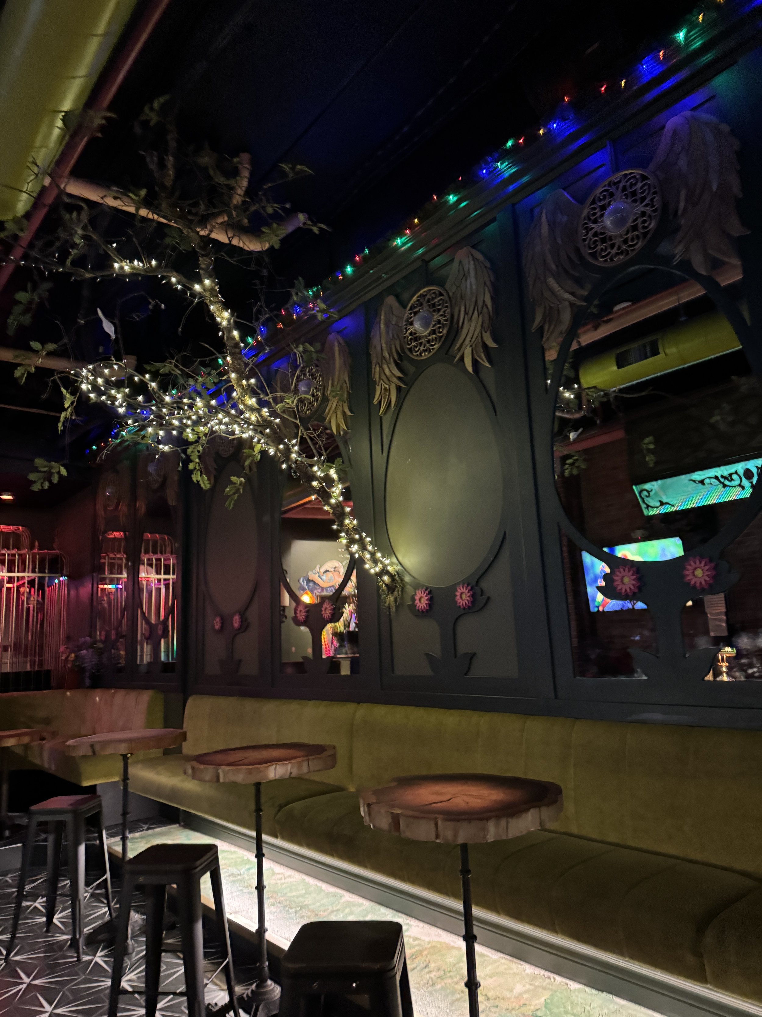 Moody bird-themed interior at Darling Aviary with winged mirrors, tree branches wrapped in lights, and green velvet seating.