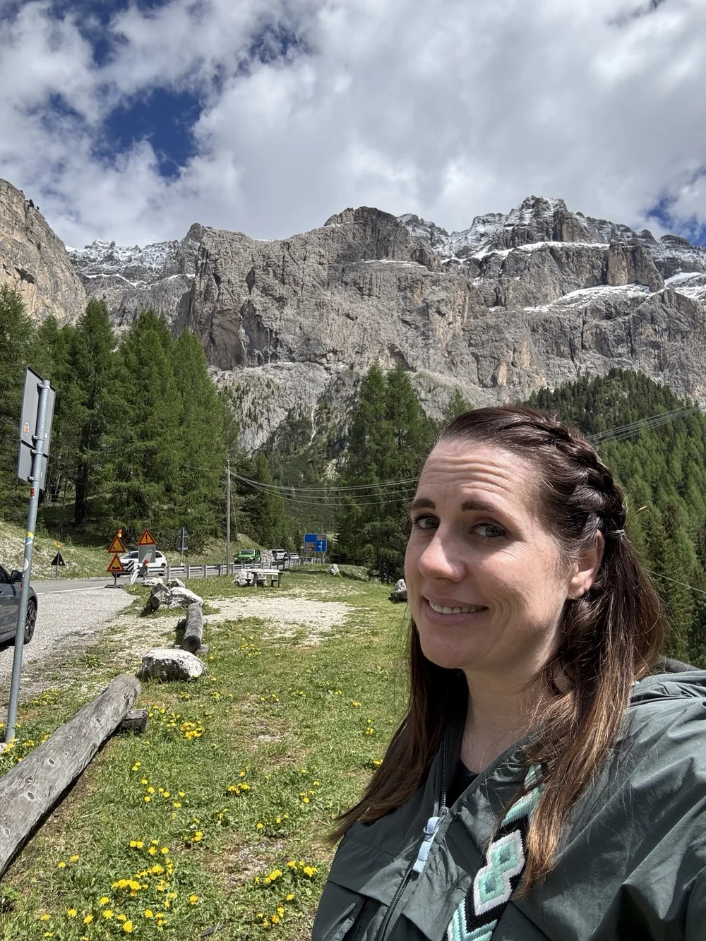 Woman taking a selfie at Sella Pass with snow-dusted Dolomite cliffs and wildflowers behind her.