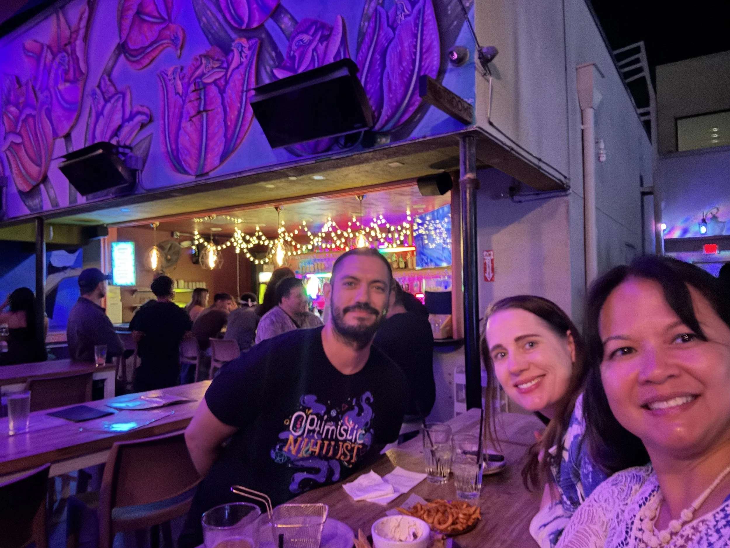 Three friends sharing food and drinks beneath colorful lighting and bird-themed artwork at Darling Aviary in Sacramento.