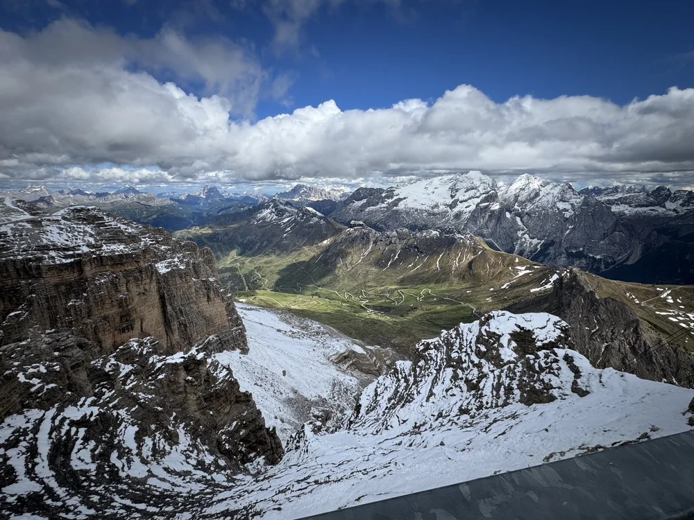 Wide view of snowy cliffs, winding mountain roads and jagged Dolomite peaks beneath a blue sky.
