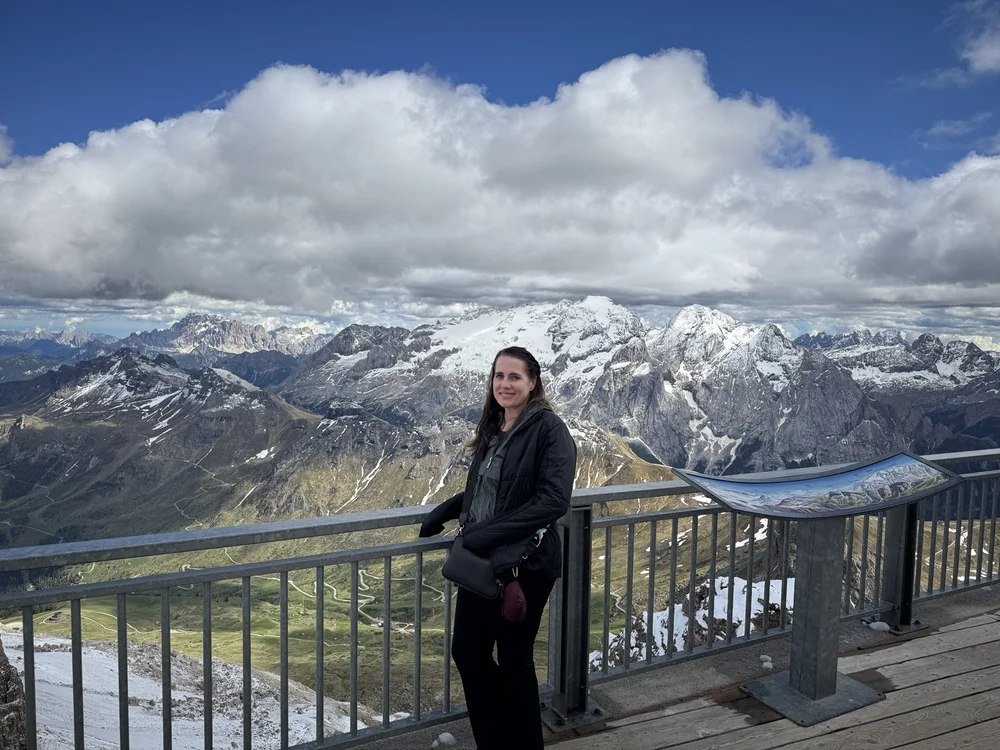 Woman standing at a Sass Pordoi viewpoint with snow-covered Dolomite mountains behind her.