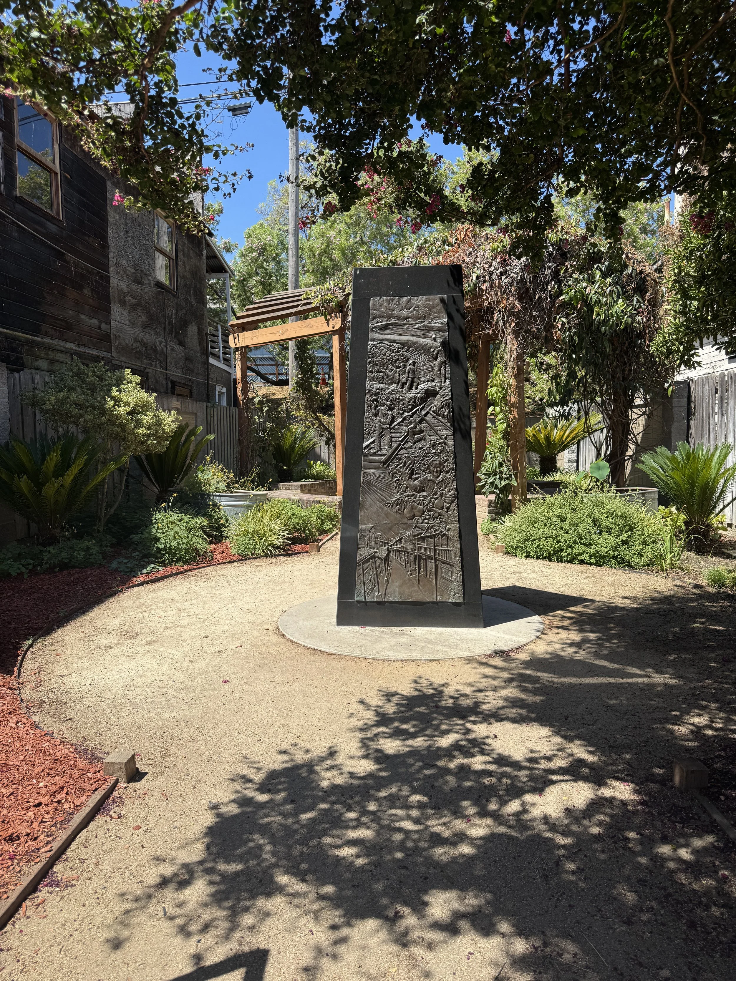 Sculpted historical monument surrounded by a shaded garden between weathered wooden buildings in Locke.