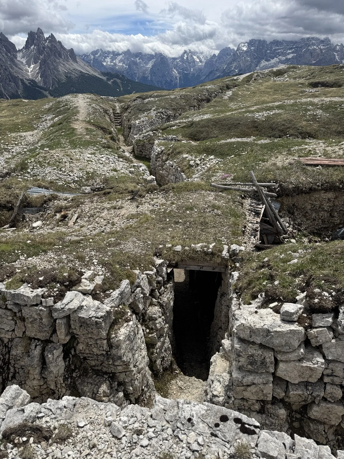 Stone-lined World War I trenches and a dark tunnel entrance with the Dolomites beyond.