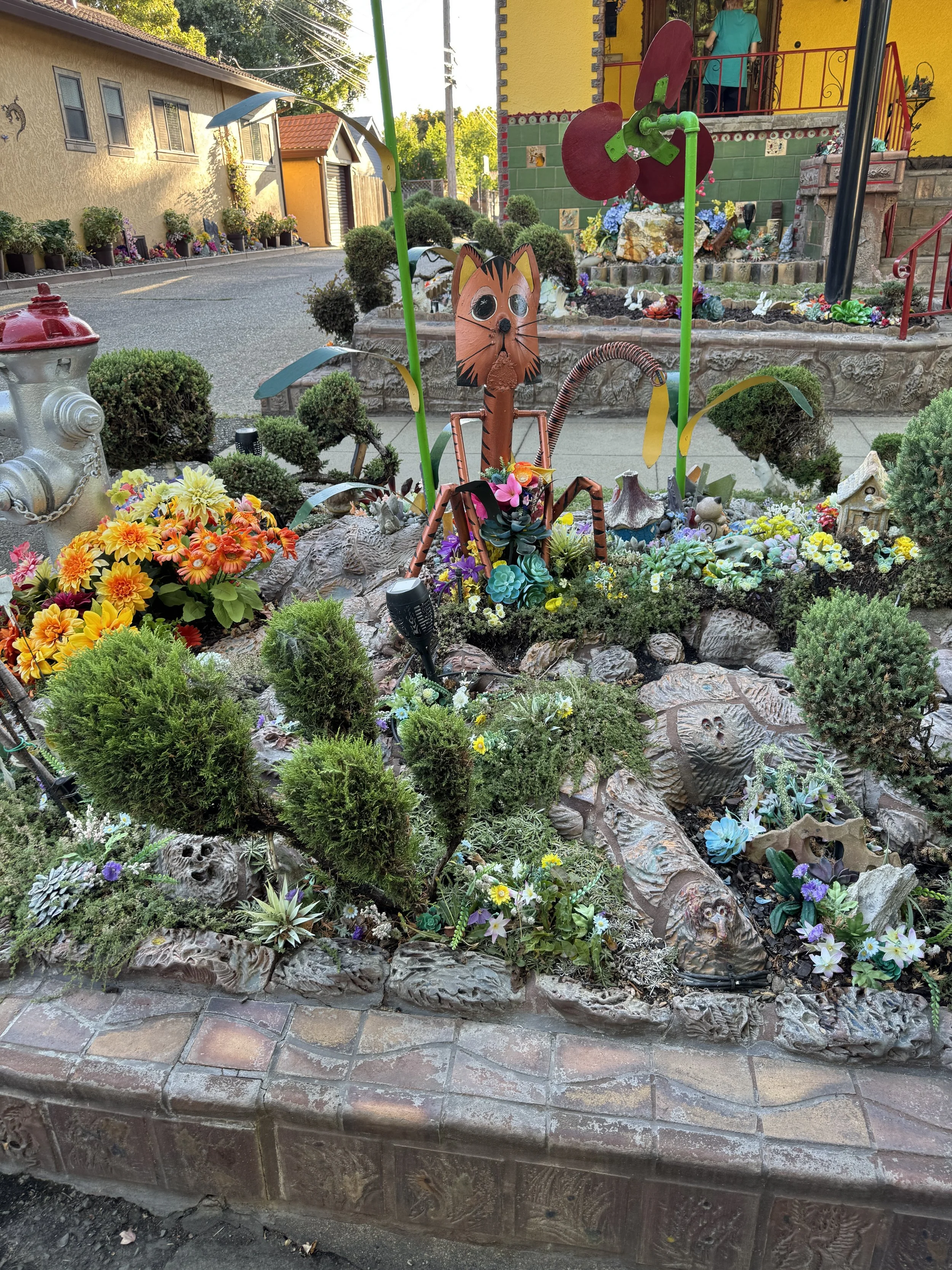 Whimsical metal cat sculpture surrounded by flowers, carved details, and miniature garden decorations at the Dragon House.