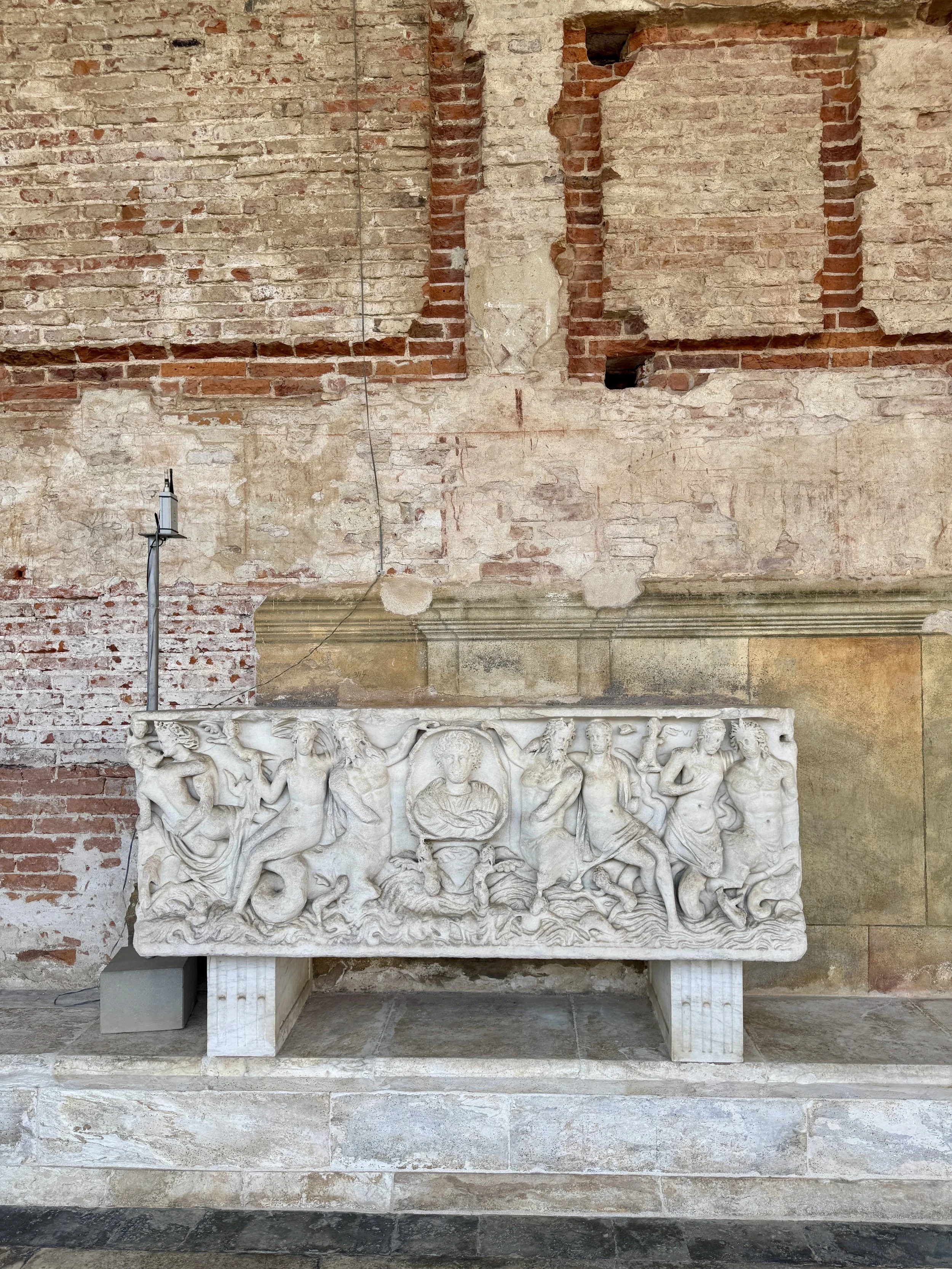 Ancient carved marble sarcophagus displayed against the brick walls of Camposanto Monumentale.