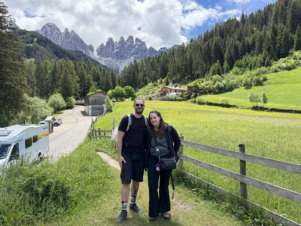 Couple standing in a green Alpine meadow with the jagged Odle peaks behind them.