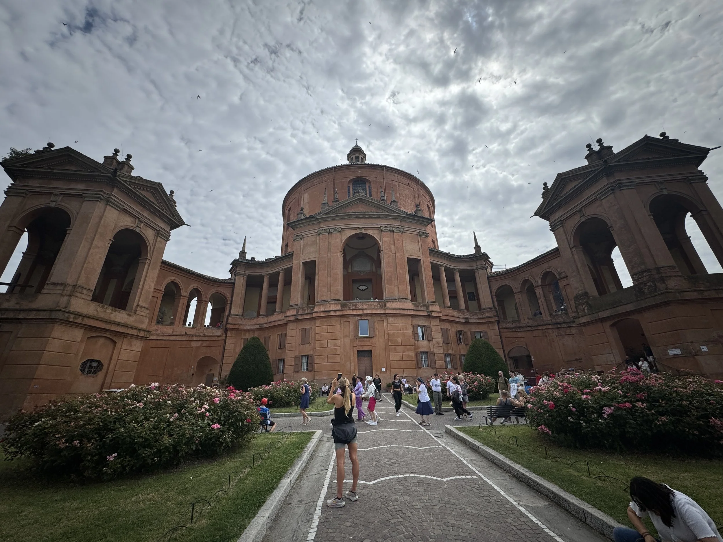 Courtyard and curved terracotta facade of the Sanctuary of the Madonna di San Luca beneath a dramatic sky.