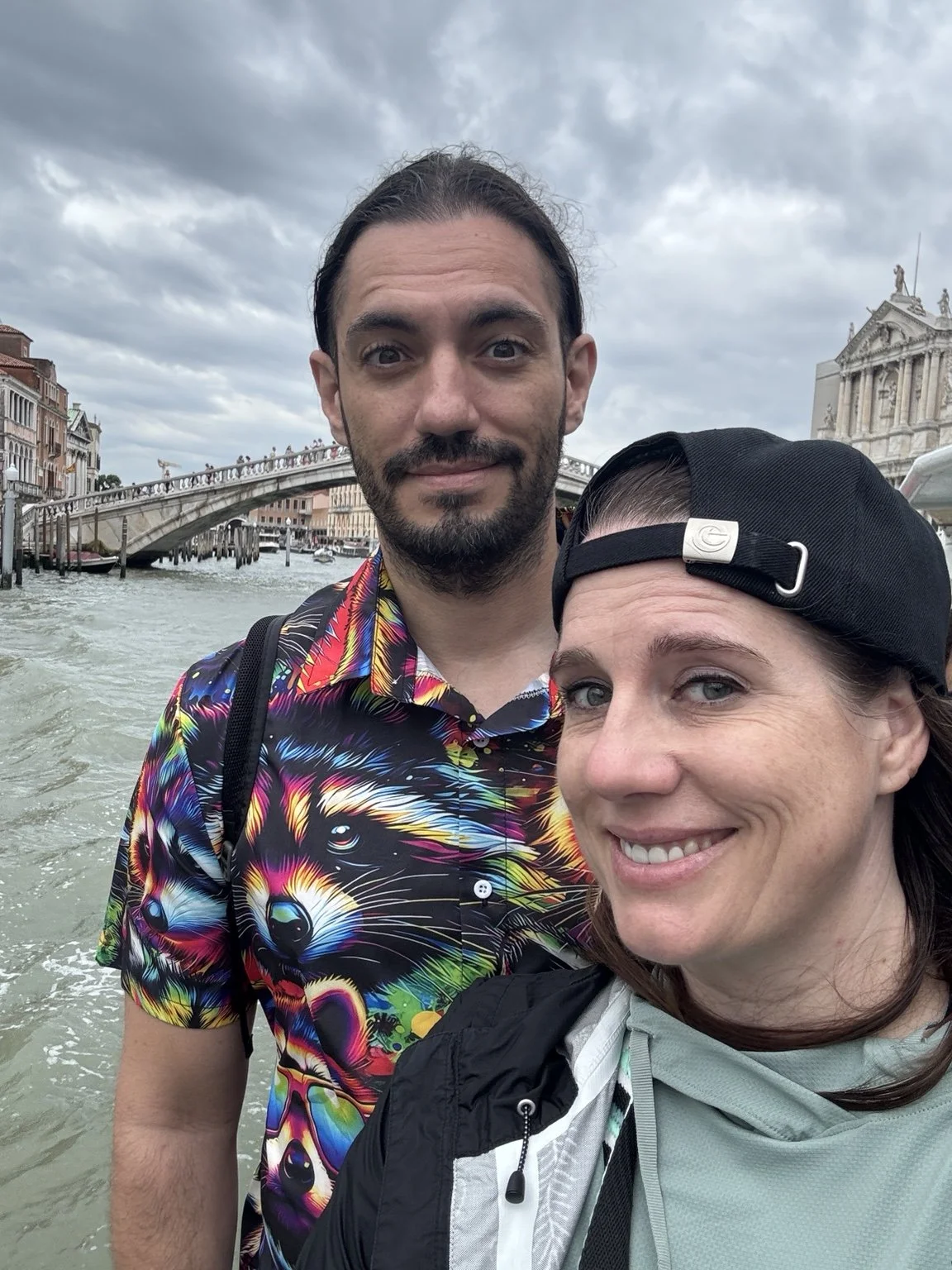 Couple taking a selfie on a Venice vaporetto with a canal bridge and historic buildings behind them.