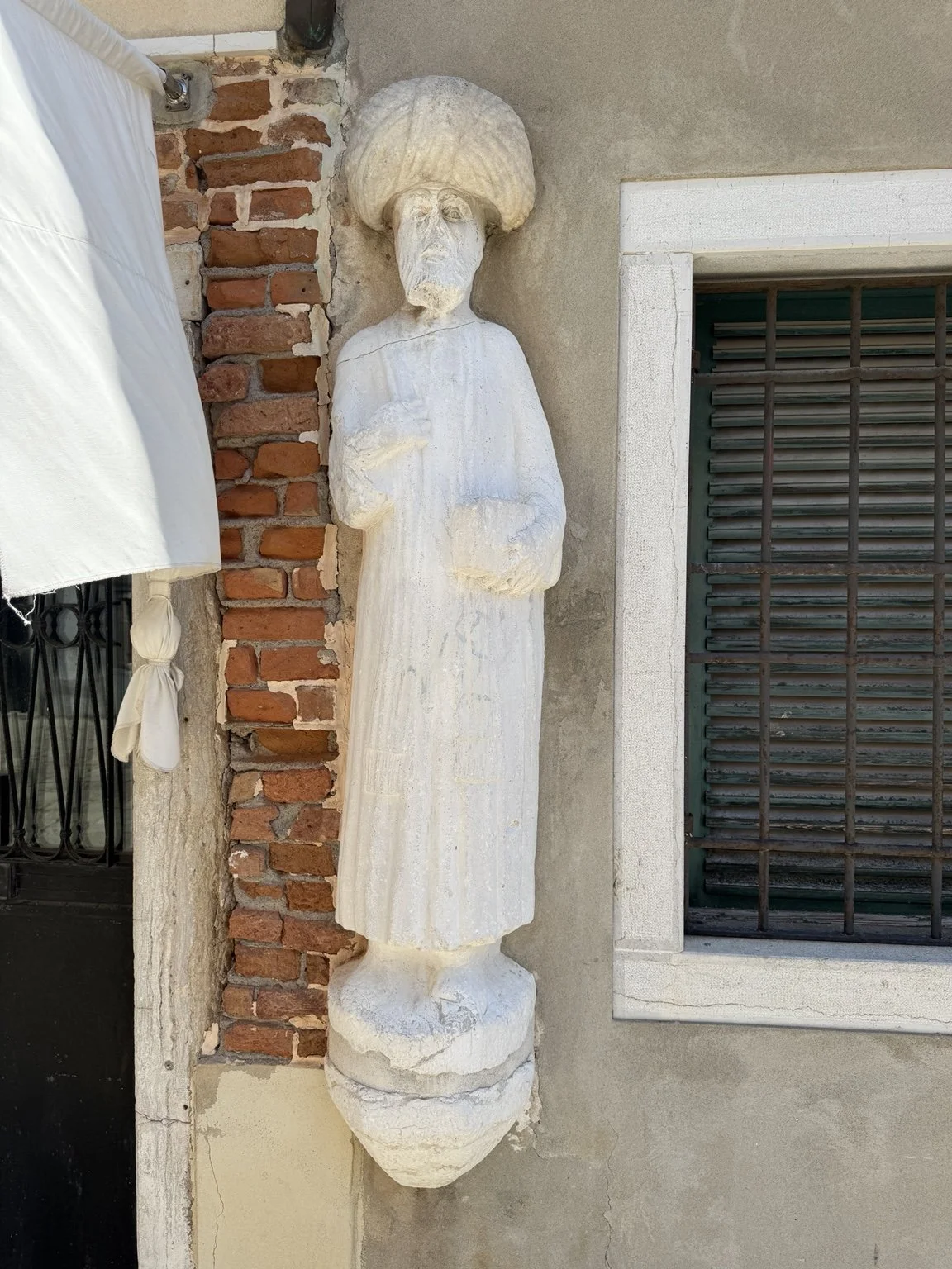 Carved stone figure from the Campo dei Mori mounted beside a shuttered window in Venice.