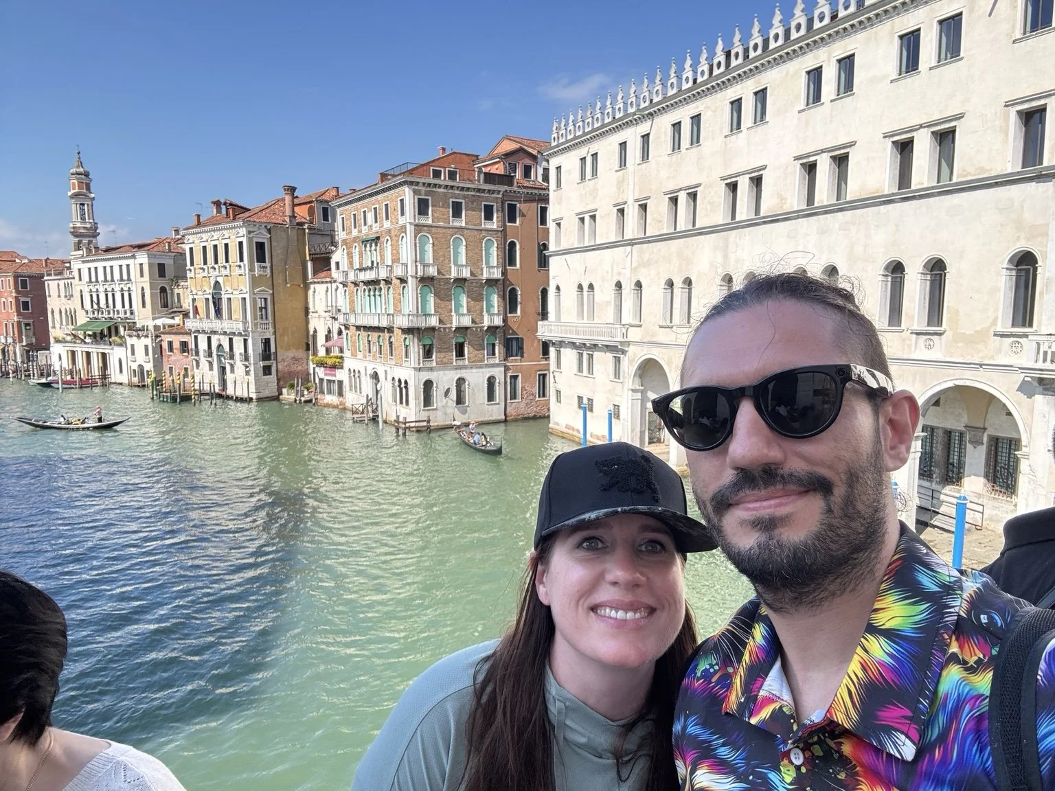Couple taking a selfie above Venice's Grand Canal with gondolas and colorful palazzi behind them.