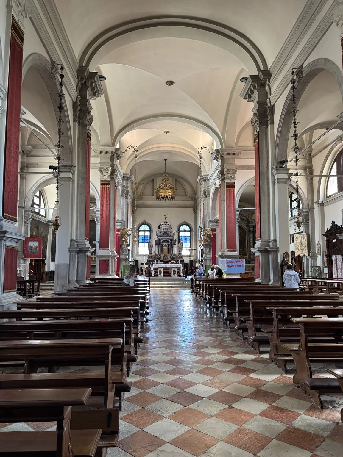 Interior of a Burano church with red columns, white arches and a checkered tile floor.