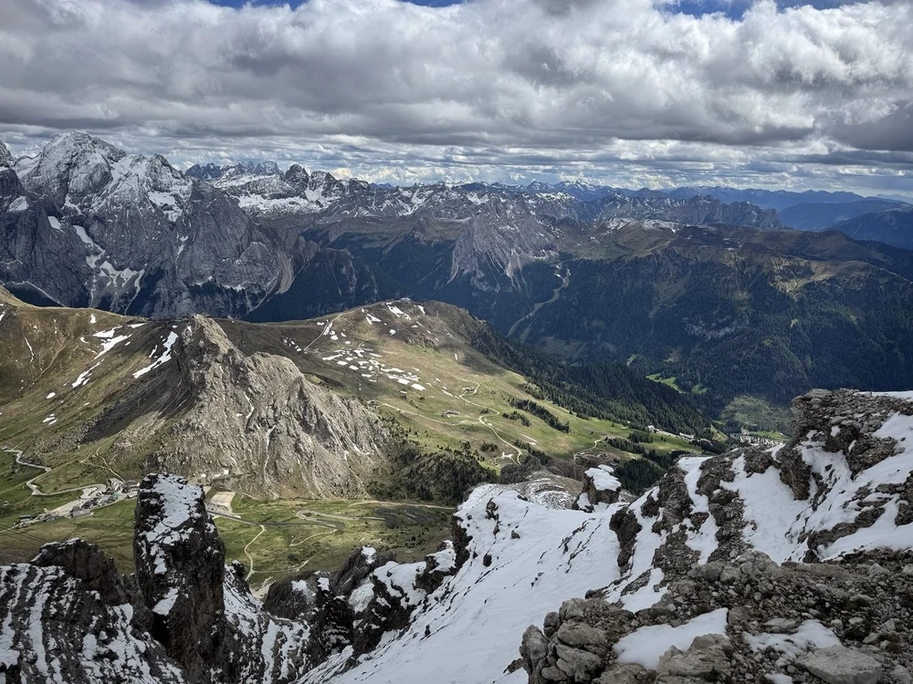 Panoramic view across snow-covered Dolomite ridges and green valleys beneath dramatic clouds.