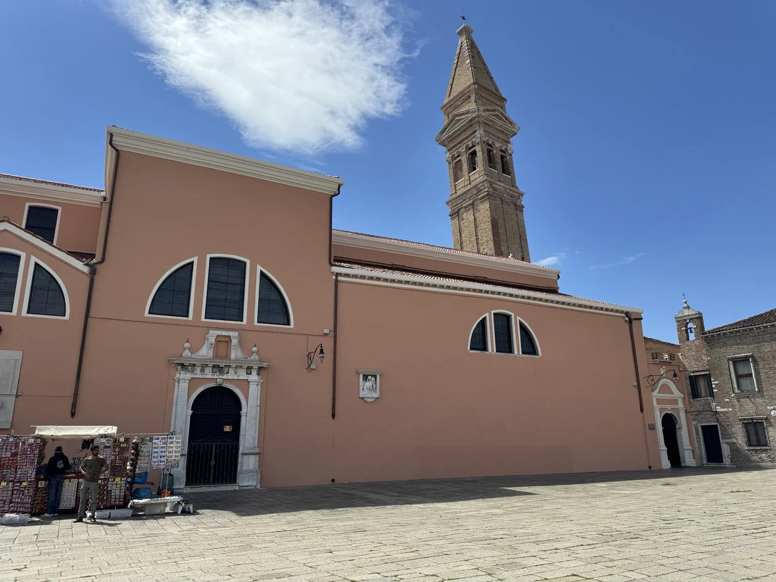 Salmon-colored church and tall brick bell tower beneath a blue sky on Burano.
