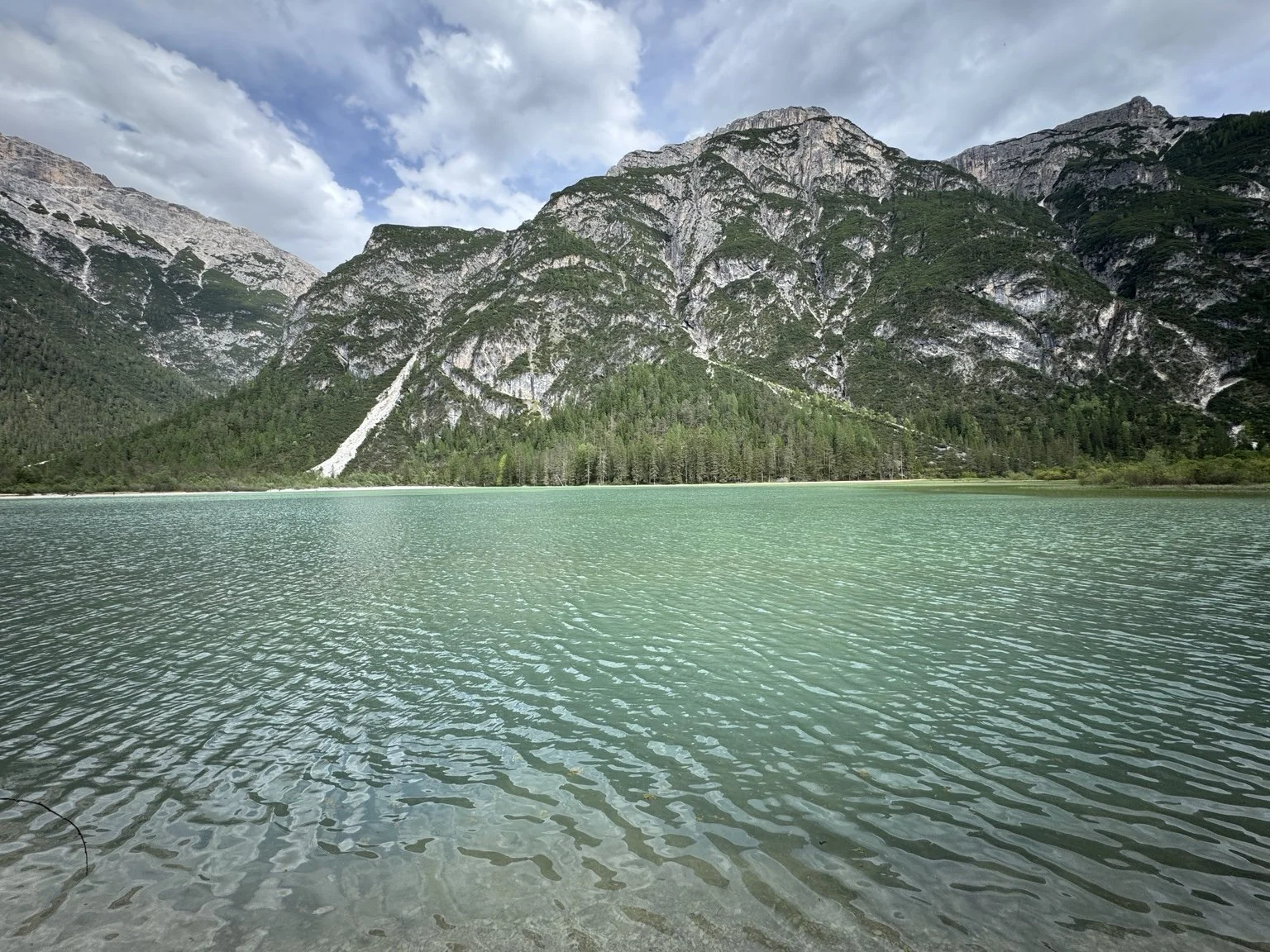 Emerald water at Lago di Landro beneath forested Dolomite mountain slopes and a cloudy sky.
