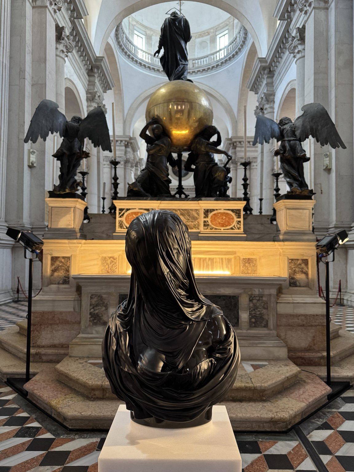 Glossy black veiled sculpture facing a gilded orb and winged figures inside San Giorgio Maggiore.