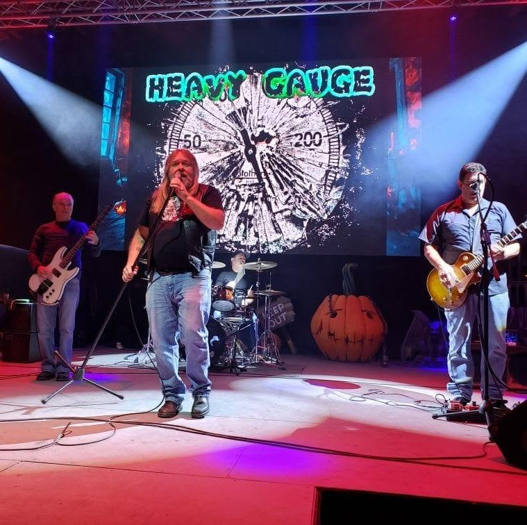 Heavy Gauge performing on stage beneath blue lights, with the band’s name and a jack-o’-lantern behind them.