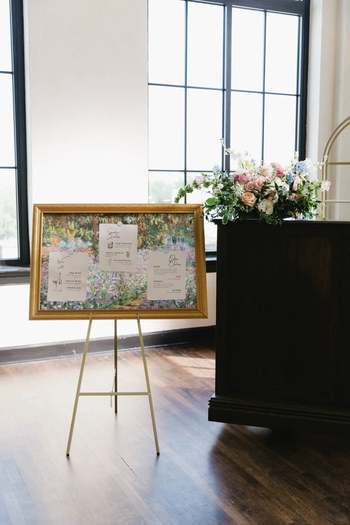 Bar arrangement of garden roses and delphinium beside a gilded menu easel