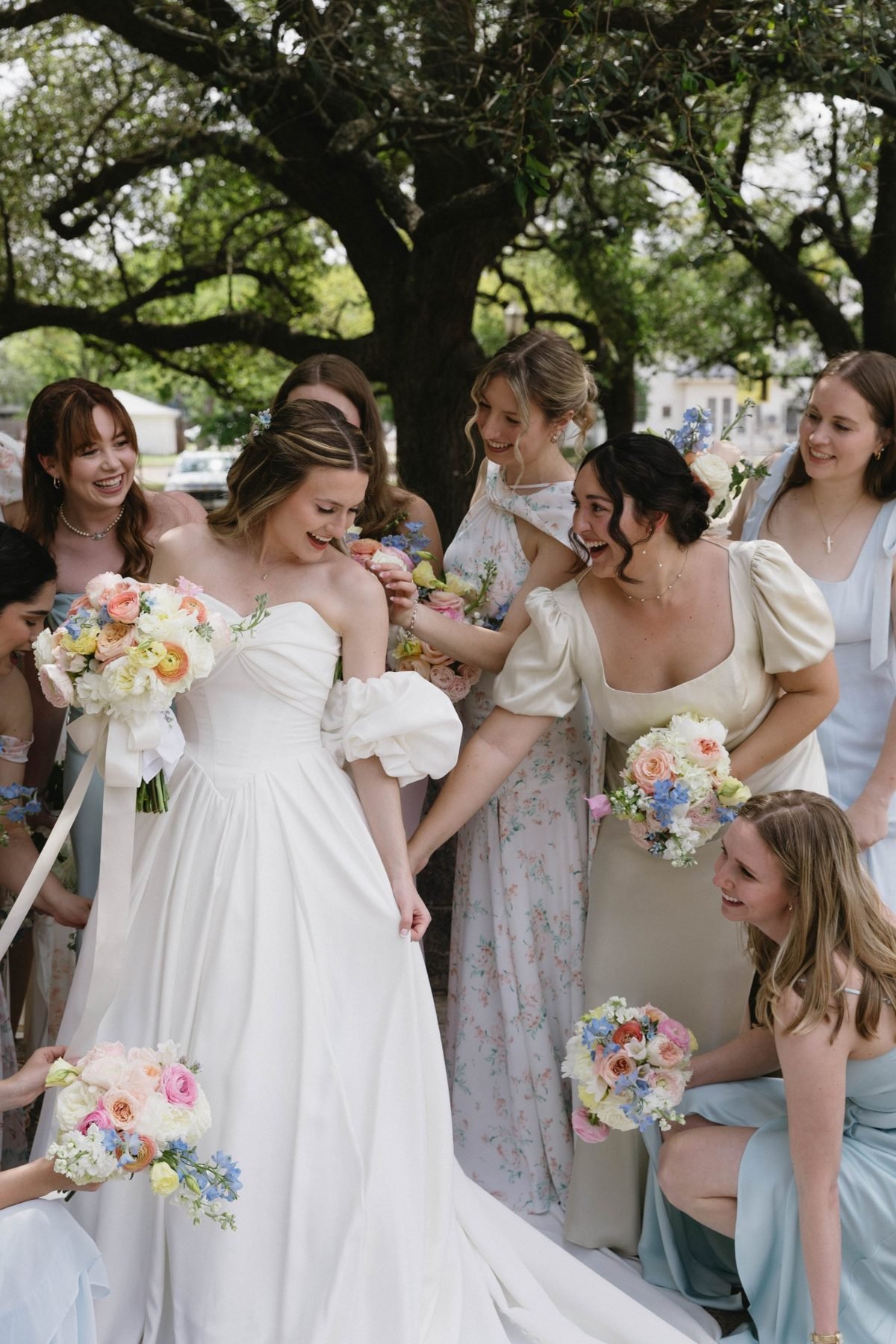 Bride and bridesmaids laughing beneath an oak tree, each holding a pastel bouquet