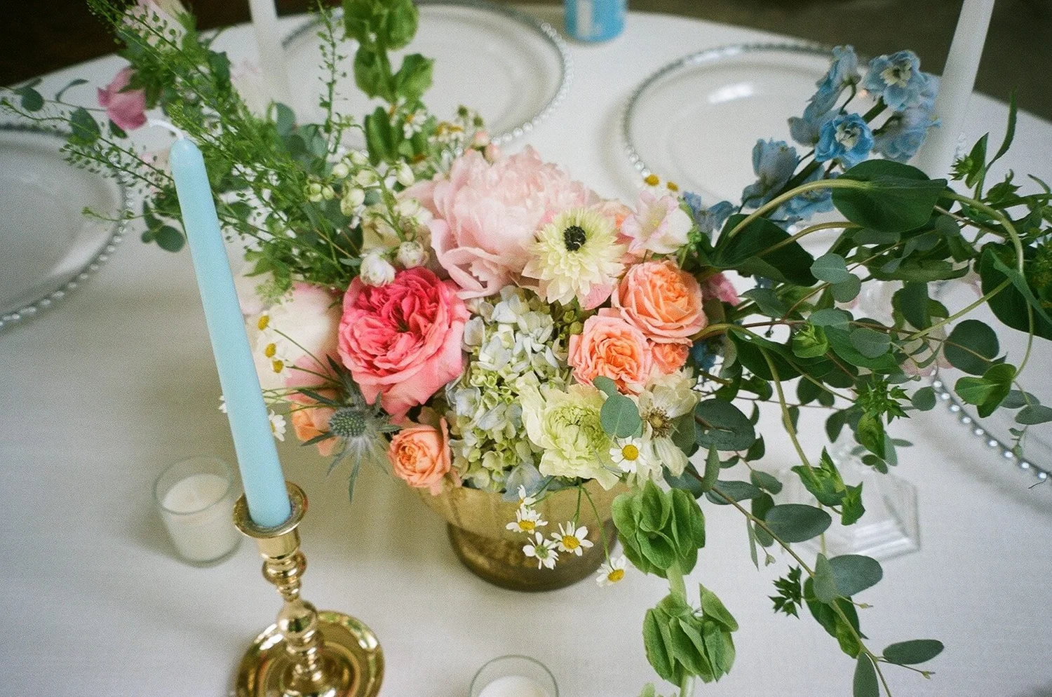 Low brass centerpiece of peony, garden roses and anemone beside a pale blue taper, shot on film