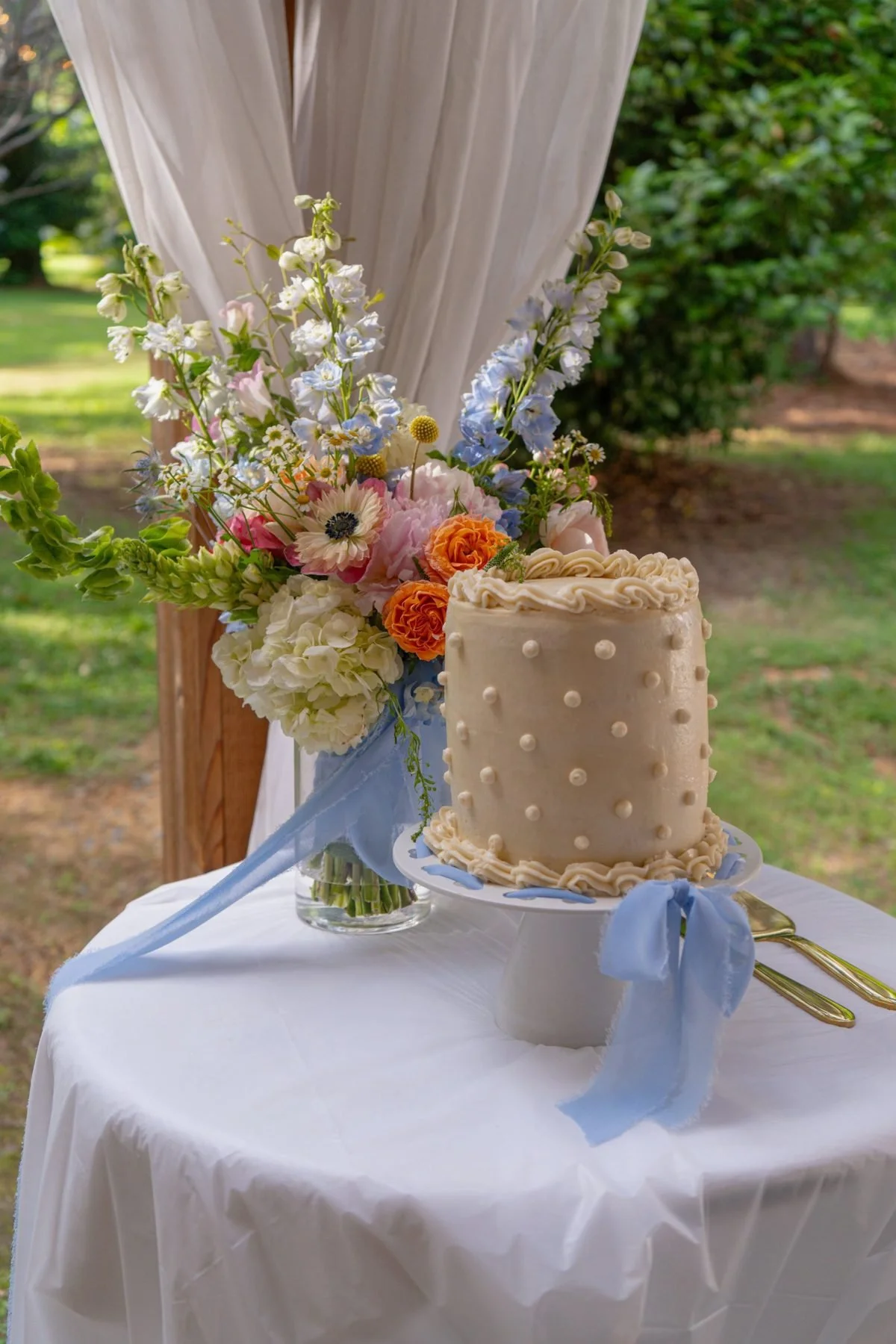 Garden arrangement of delphinium, peonies and anemones beside a polka-dot cake tied with blue silk