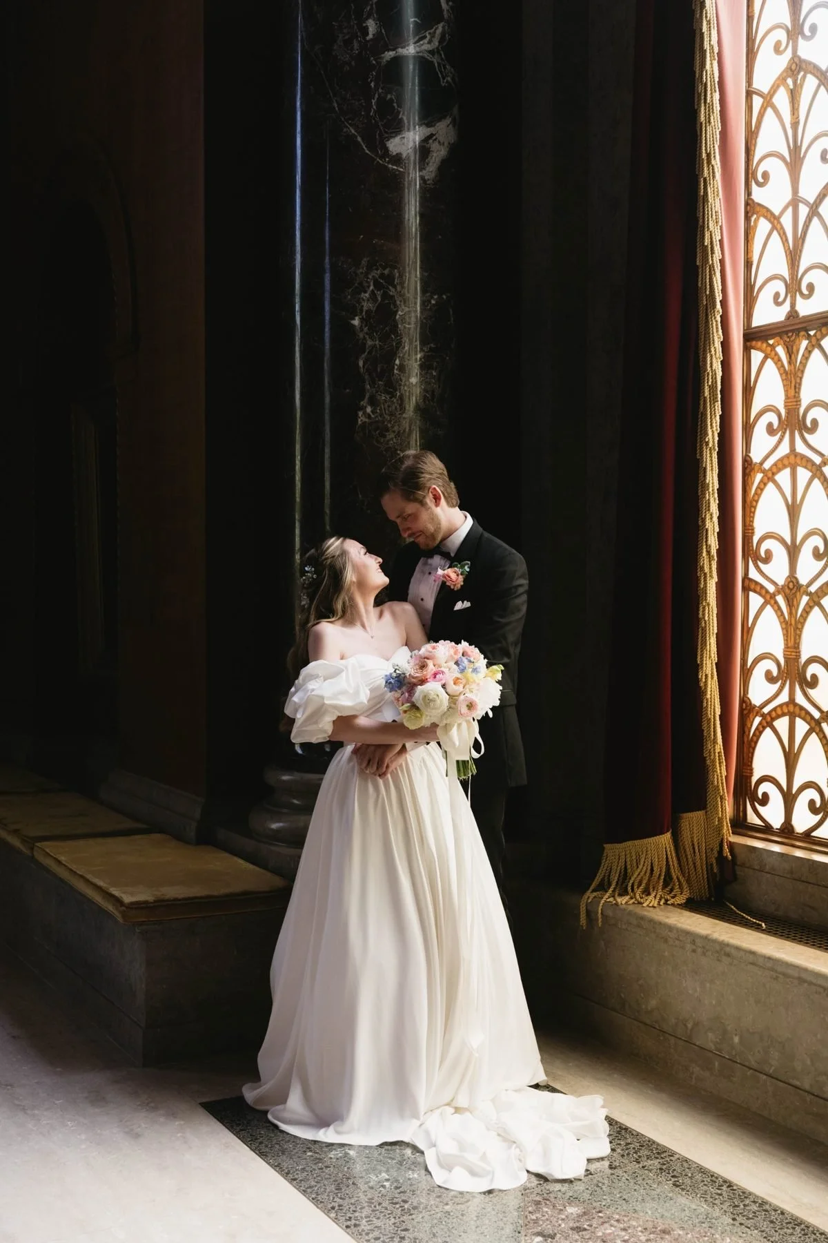 Couple embracing beside a gilded window, bouquet held between them