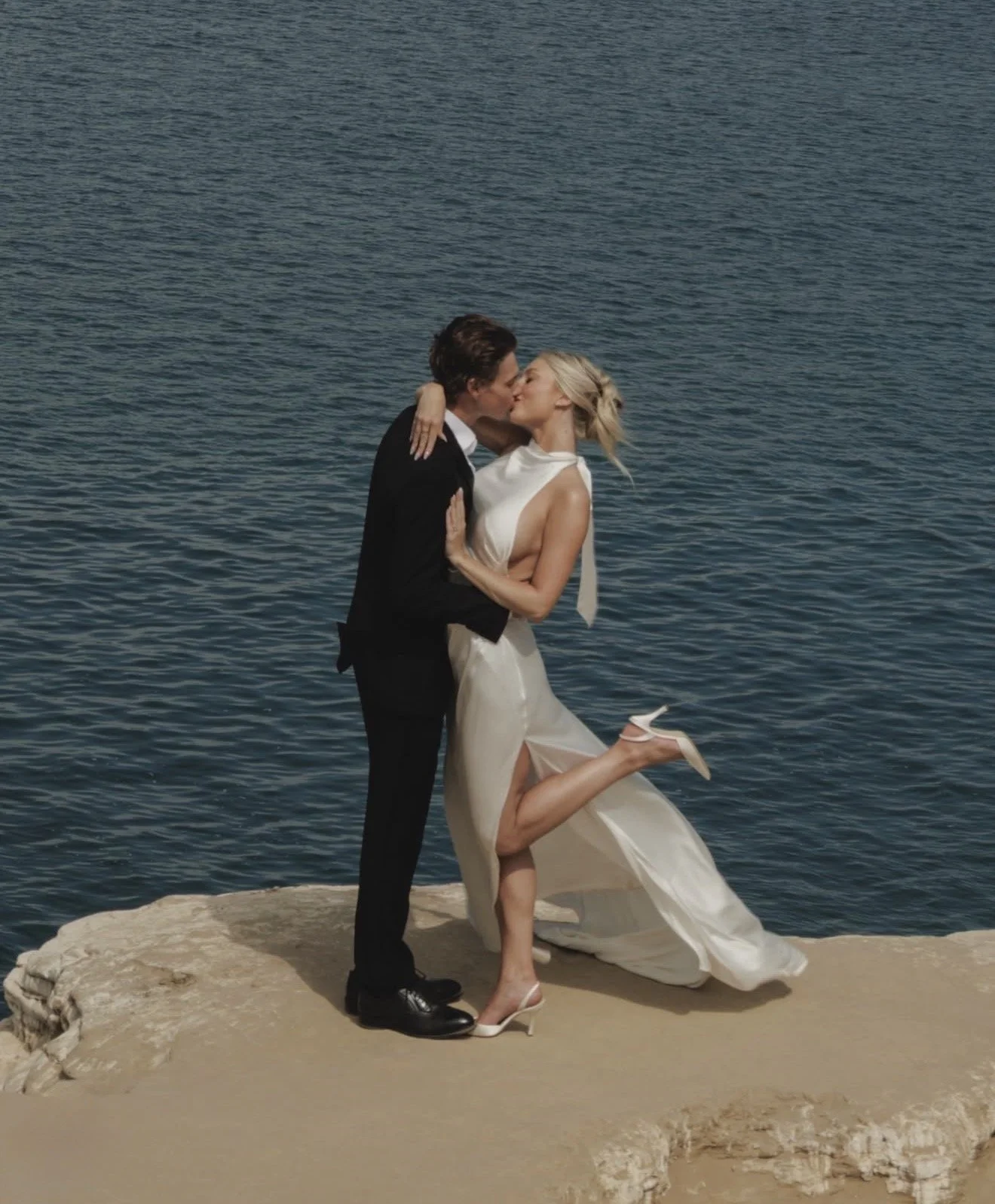Newlyweds kissing on a rocky shoreline beside blue water