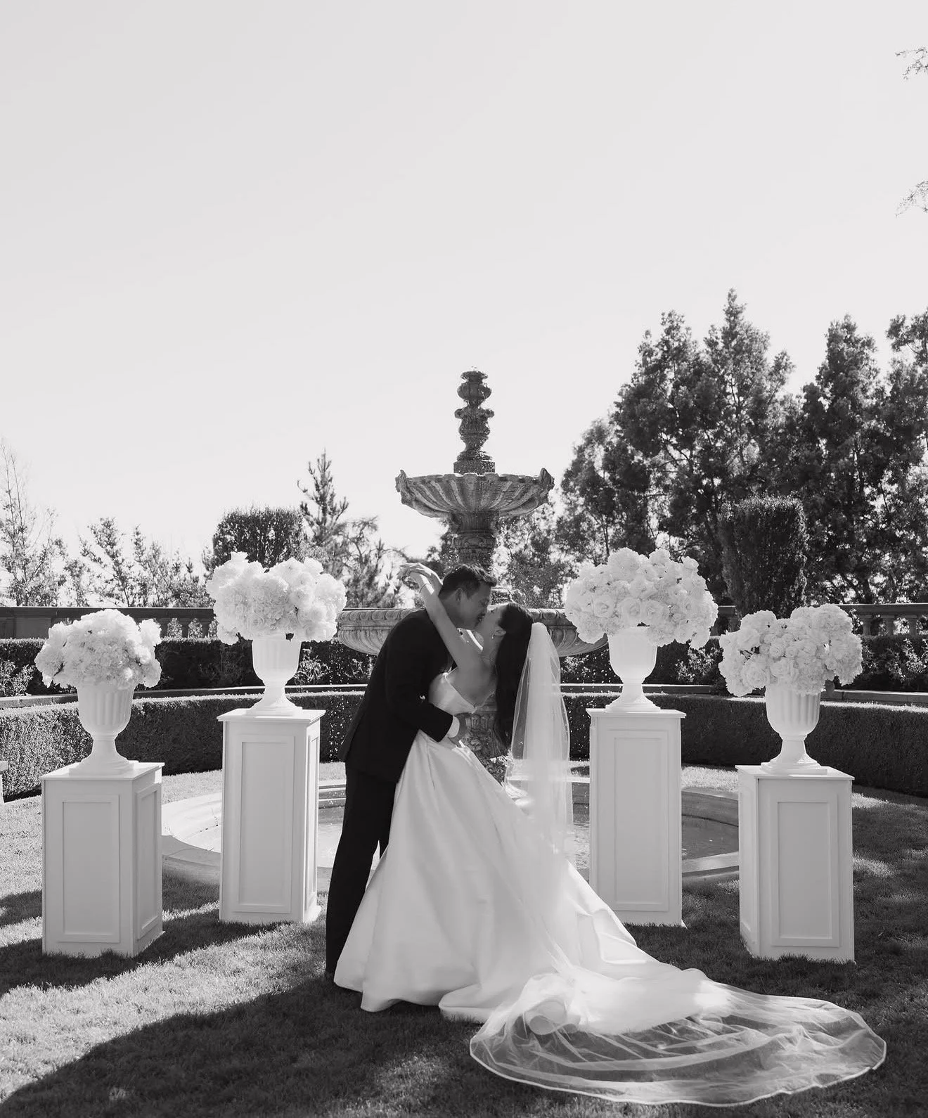 Black-and-white wedding portrait beside a fountain and white floral urns