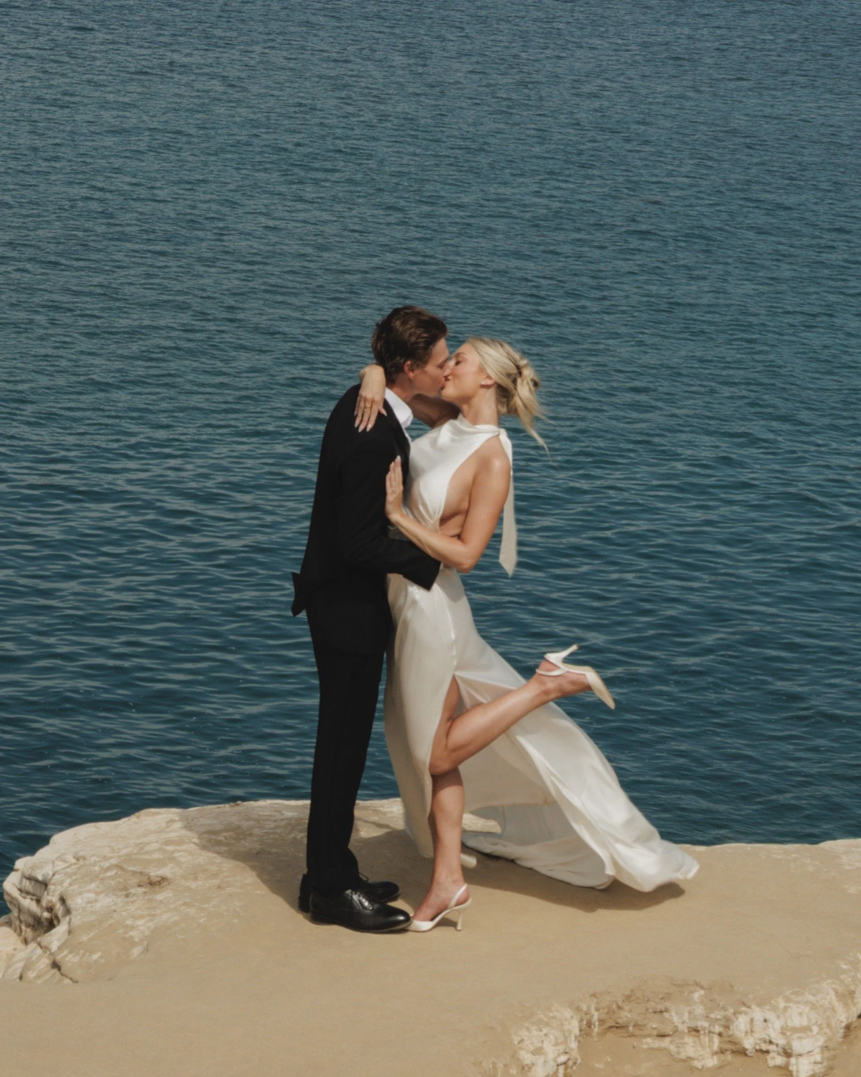 Newlyweds kissing on a rocky shoreline above blue water