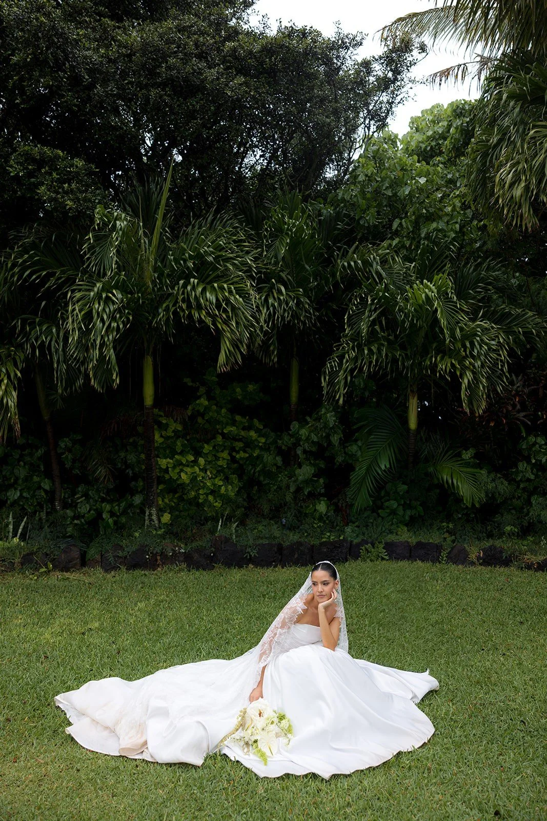 Bride seated on grass beside tropical greenery