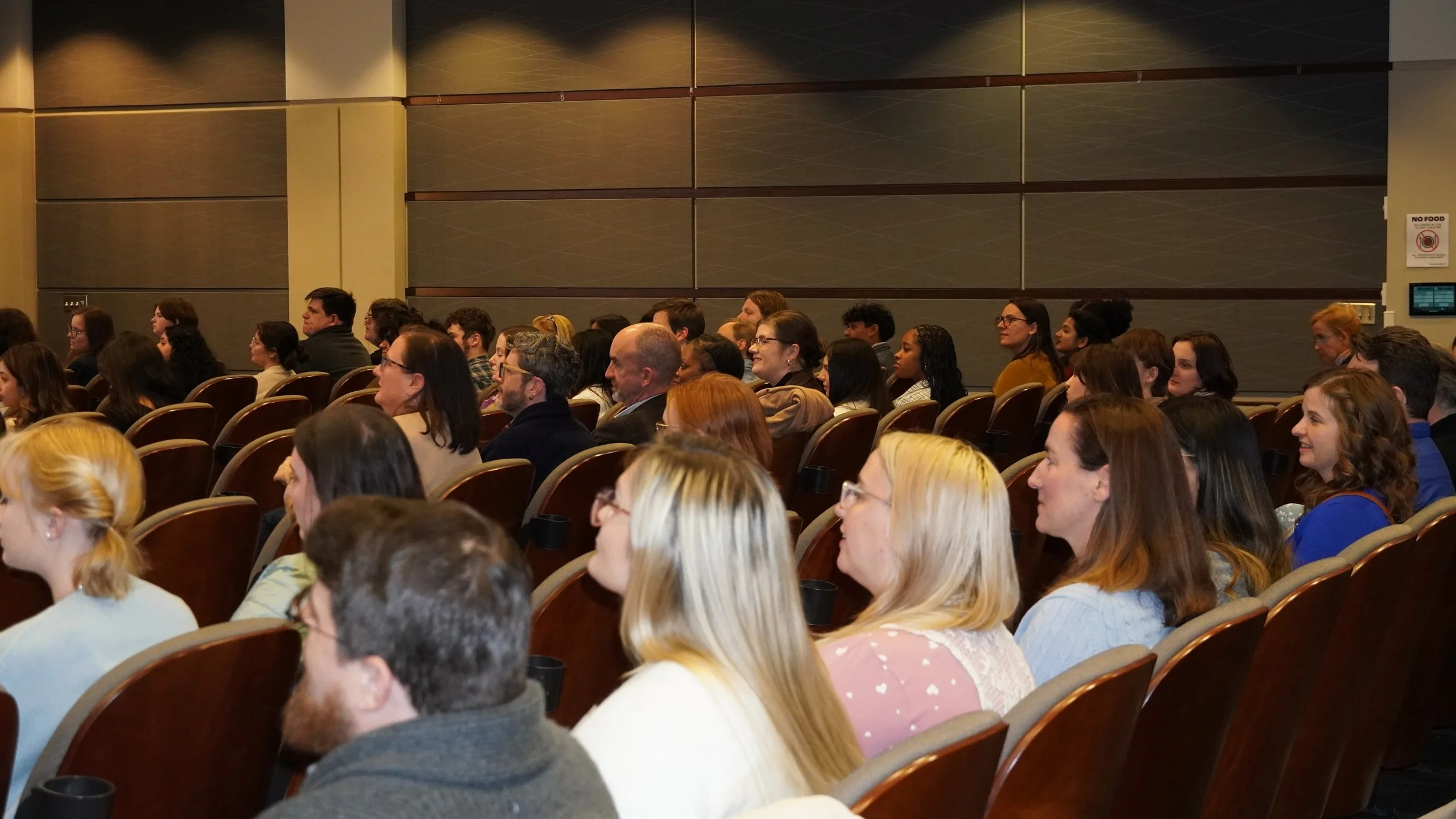 An auditorium of people listen to a presentation with their backs turned towards the camera.