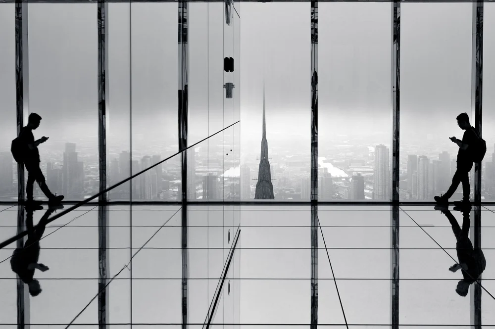 Black and white: two silhouetted men on phones stand at floor-to-ceiling glass with their reflections in the mirrored floor, the Chrysler Building's spire dead centre in flat white haze.