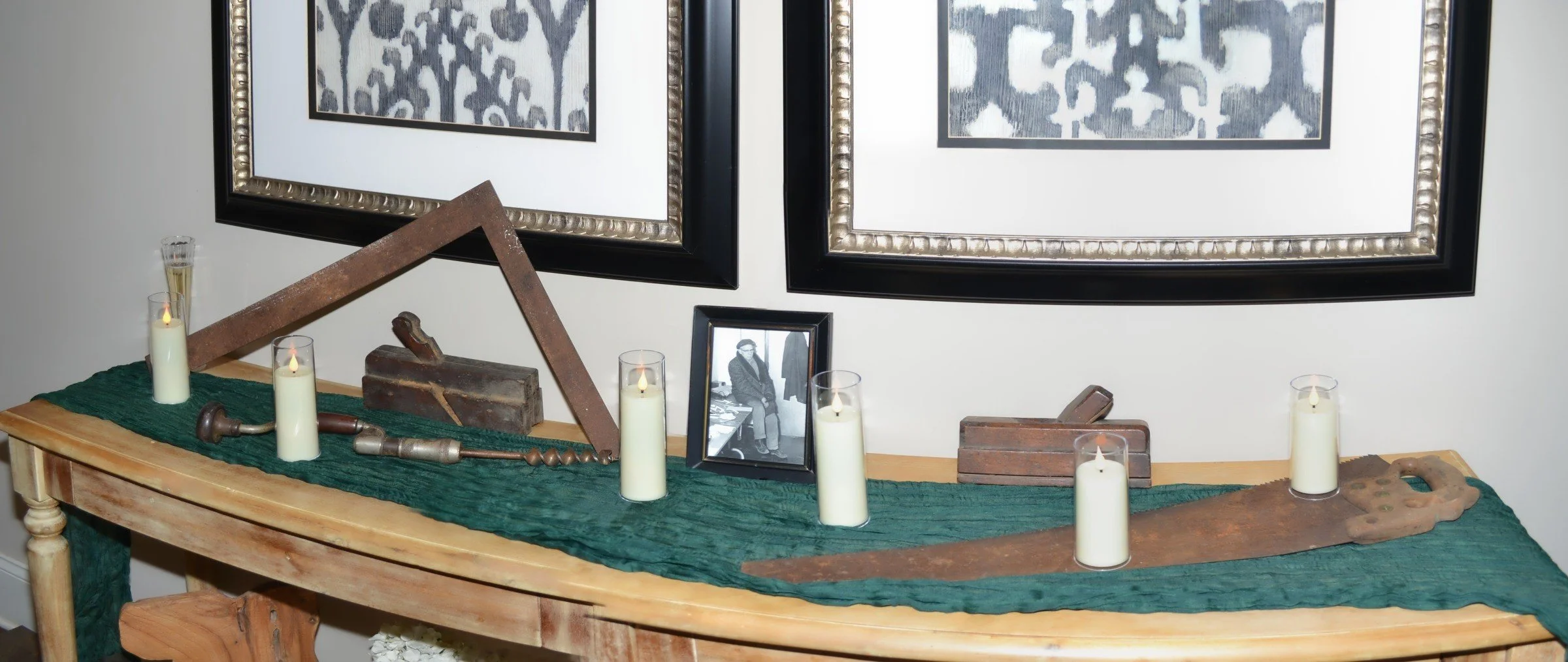 The complete memorial tribute table for Miles Murtha O'Brien Sr. — framing square, molding planes, hand brace, framed photograph, and antique handsaw, arranged on the green runner beneath the ikat prints