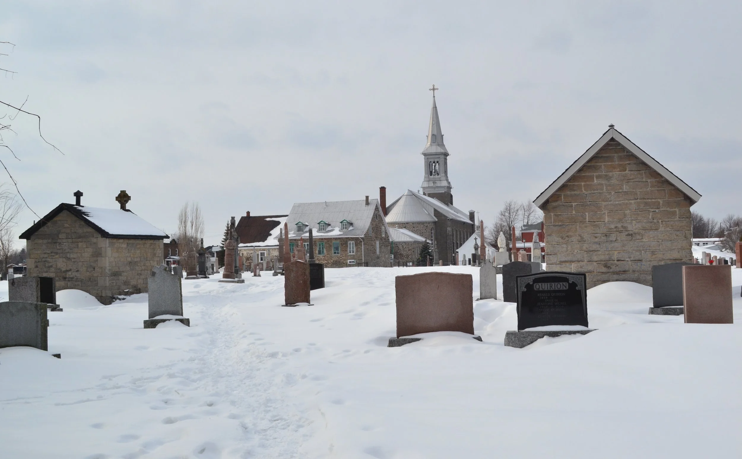 The old presbyteries of Saint-Constant in winter — two stone buildings flanking a snow-covered cemetery, headstones in the foreground, the parish church and steeple behind