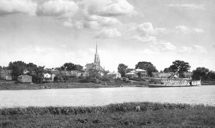 Photographie du village de Contrecoeur vers 1915, vue des îles, avec le bateau à vapeur Terrebonne accosté au quai et l'église Sainte-Trinité dominant le village