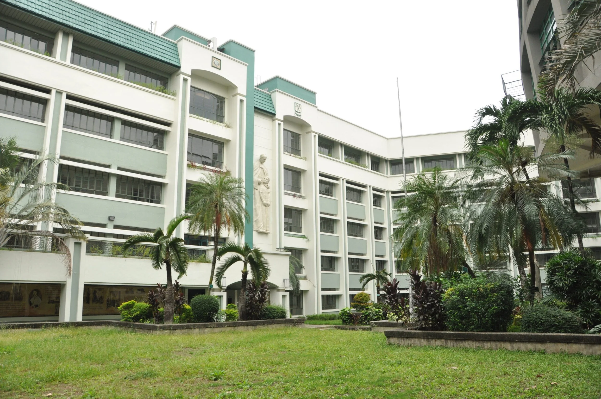The green-roofed academic buildings of La Salle Green Hills, with the relief sculpture of St. John Baptist de La Salle on the facade, framed by palm trees and lawn