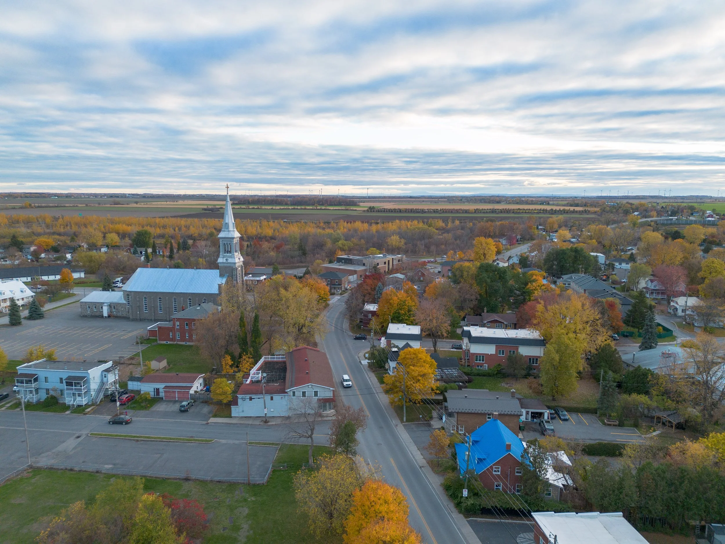 Aerial view of Saint-Constant, Quebec in autumn — the parish church and its steeple at left, the main road running through the village, farmland stretching to the horizon