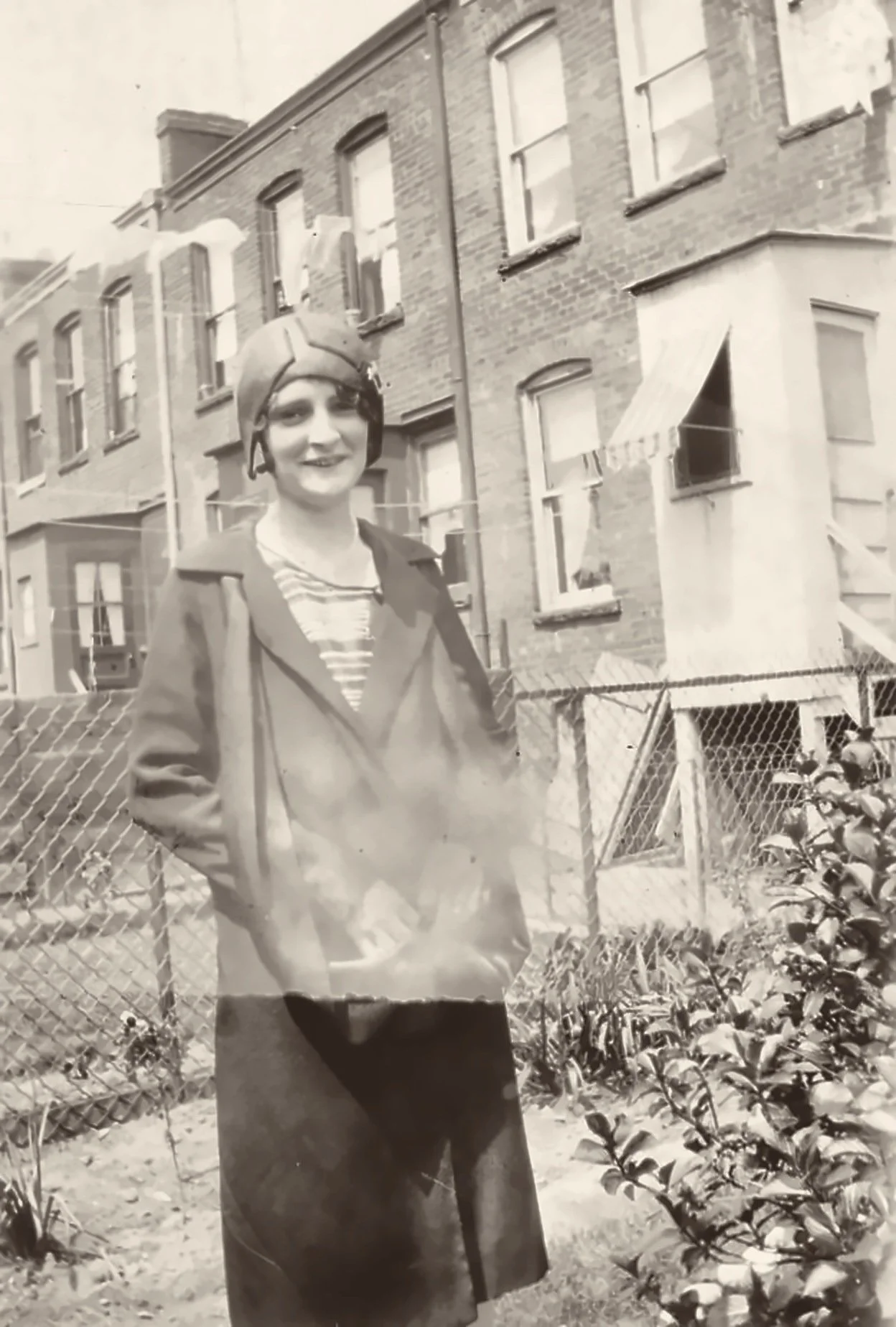 Lillian Robertson O'Brien in a brimless paneled helmet cloche and long wrap coat over a striped top, standing in a Brooklyn backyard before brick rowhouses with a clothesline and striped awning, May 1929