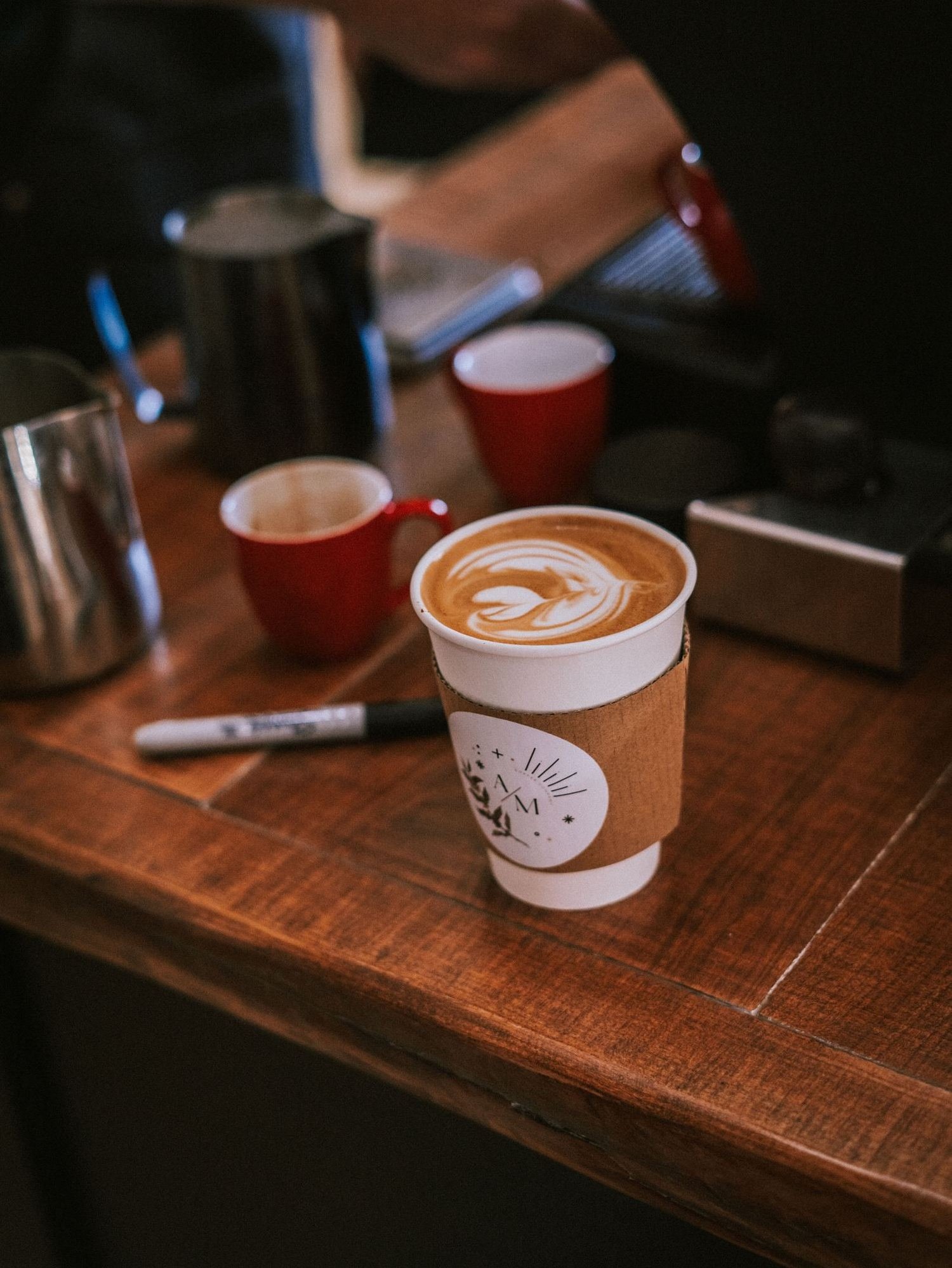 Latte in an AM Coffee Co branded cup on a wood bar beside red espresso cups