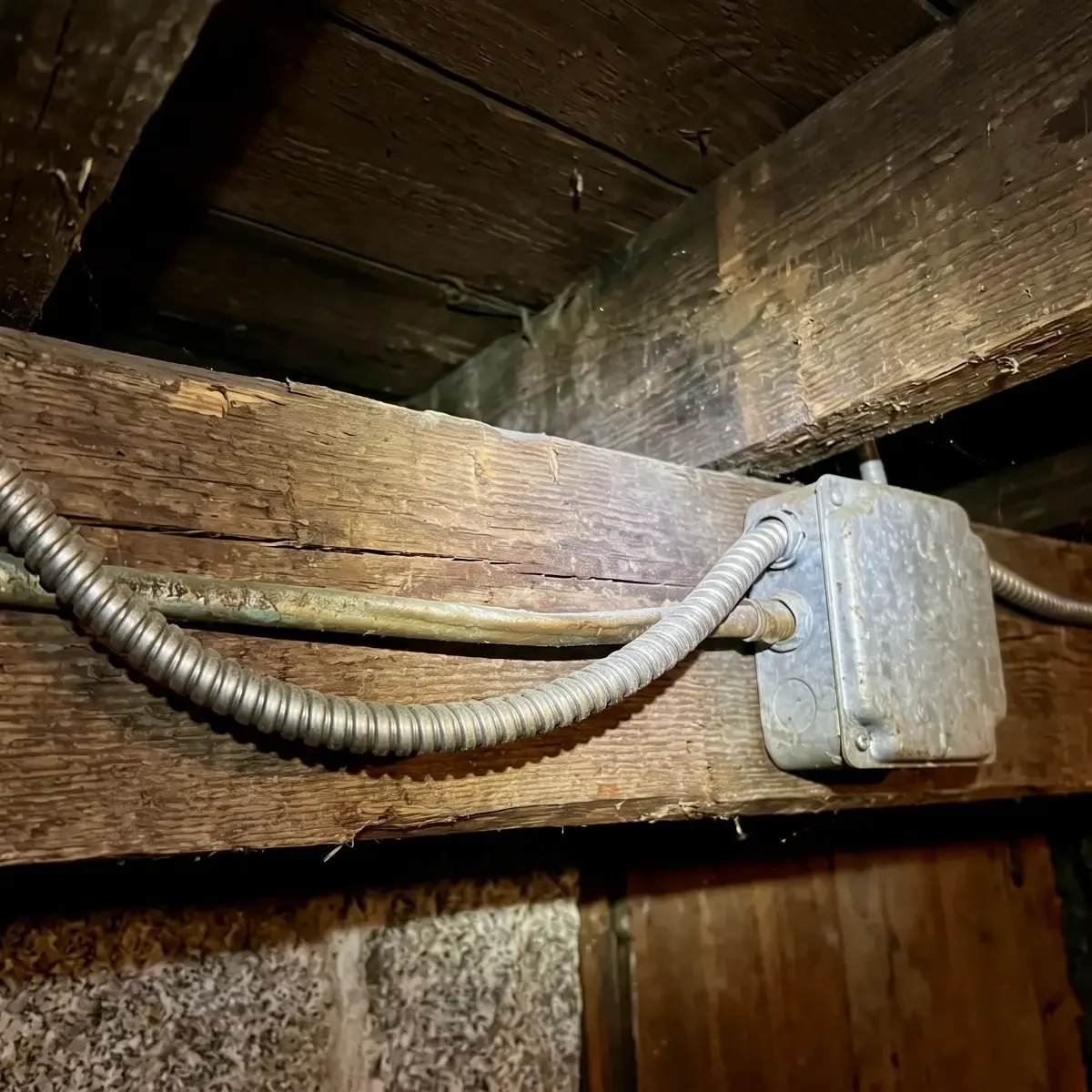 Old BX armored cable with its spiral metal jacket running along a wooden basement beam into a metal junction box.
