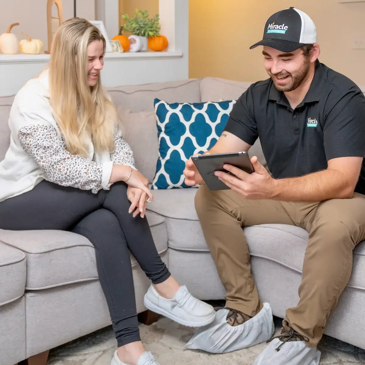Parker Gammon of Miracle Electric, in a branded cap and boot covers, sits on a homeowner's couch and shows her the results of the safety check on a tablet.