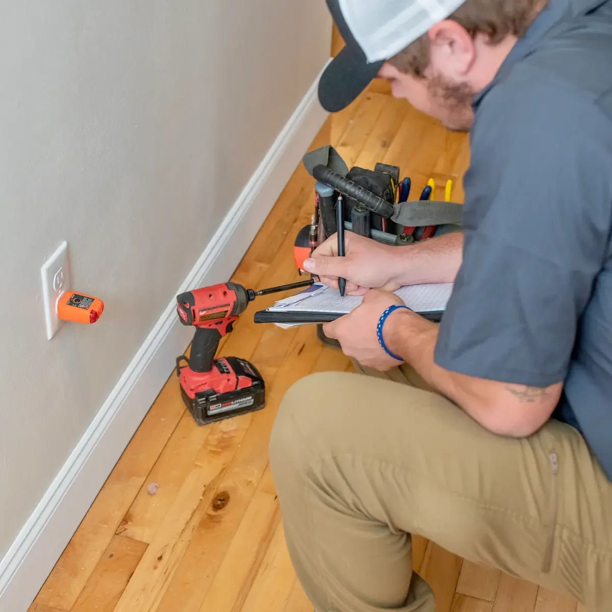 Miracle Electric electrician kneeling beside a wall outlet with a plug-in outlet tester, writing down results on a clipboard.