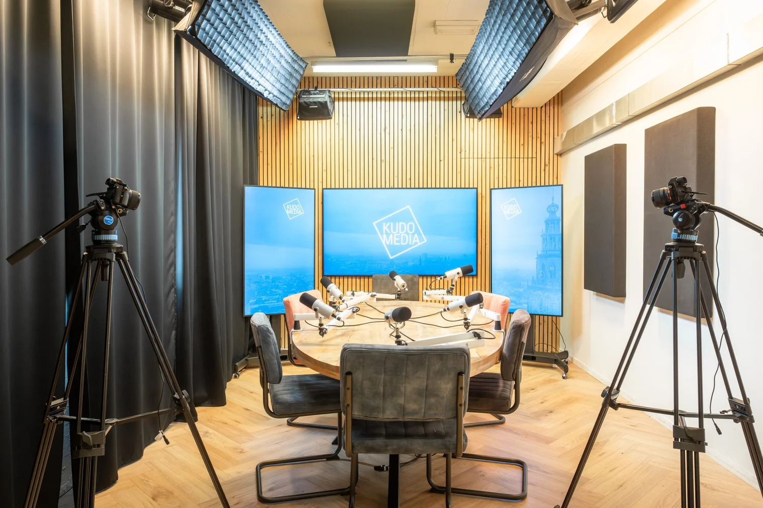 Volledige opstelling van de podcaststudio van Kudo Media in Groningen met ronde tafel, microfoons en drie 4K-schermen
