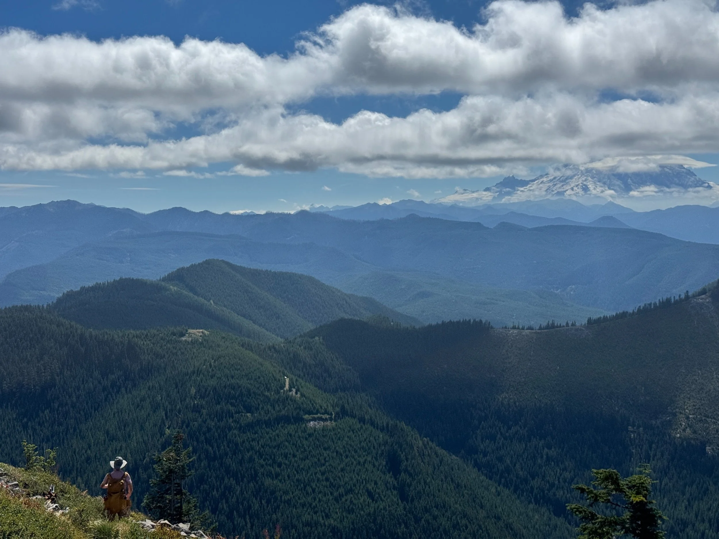 Expansive Cascade landscape with cloud-wrapped Mount Rainier and a small person at lower left.